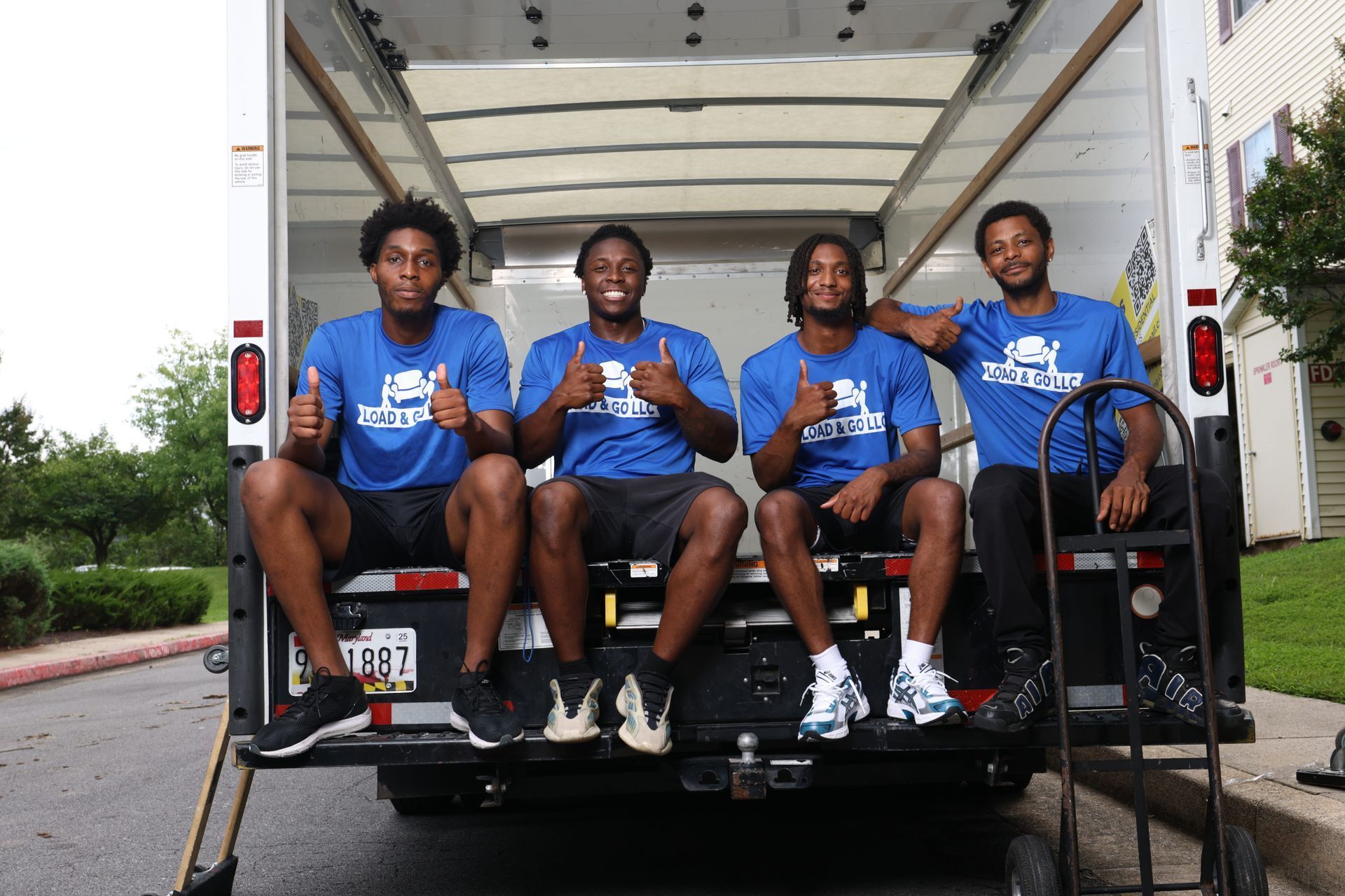 Four people in blue shirts sit on the back of a white truck, smiling outdoors.