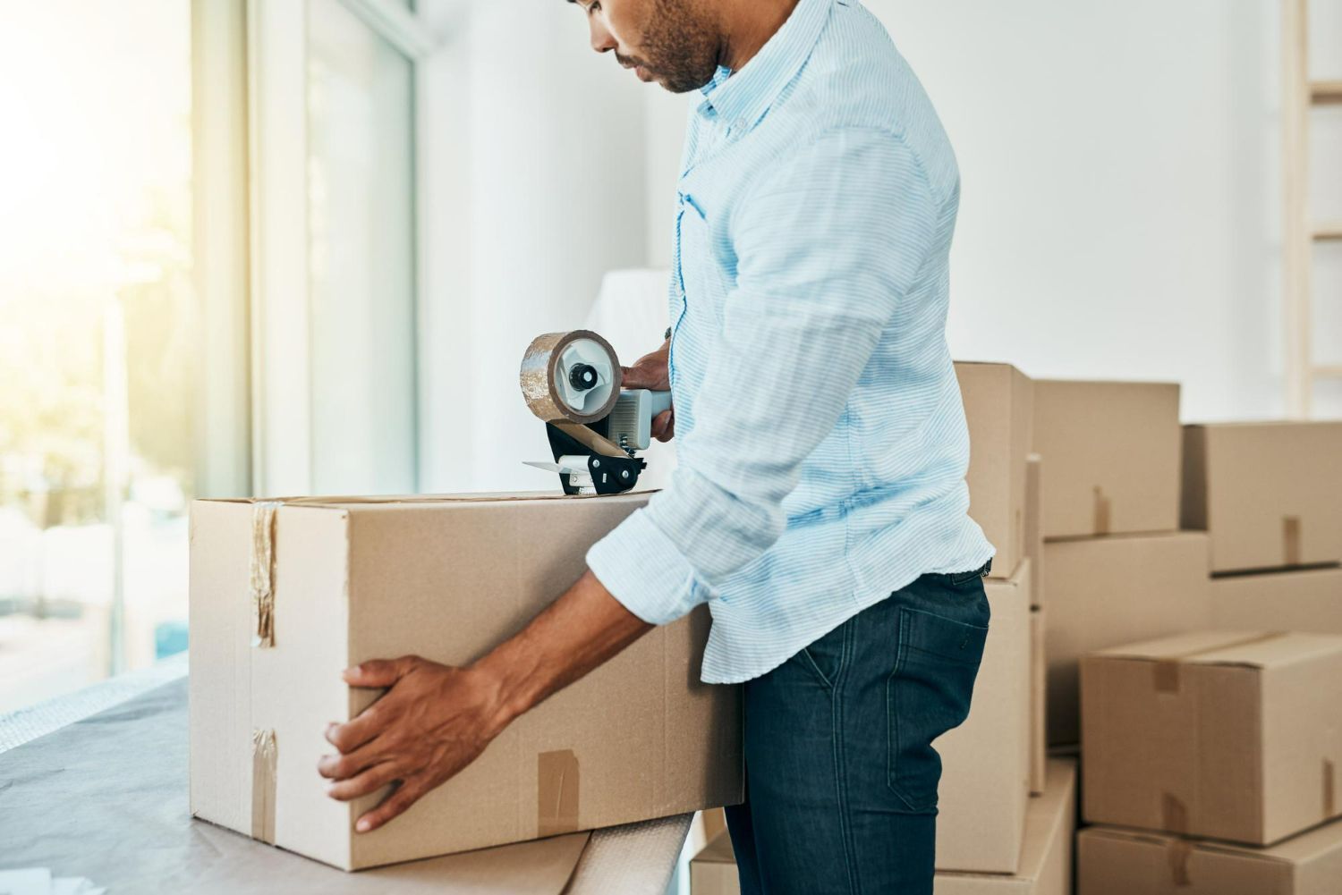 Person sealing a cardboard box with packing tape in a bright moving room