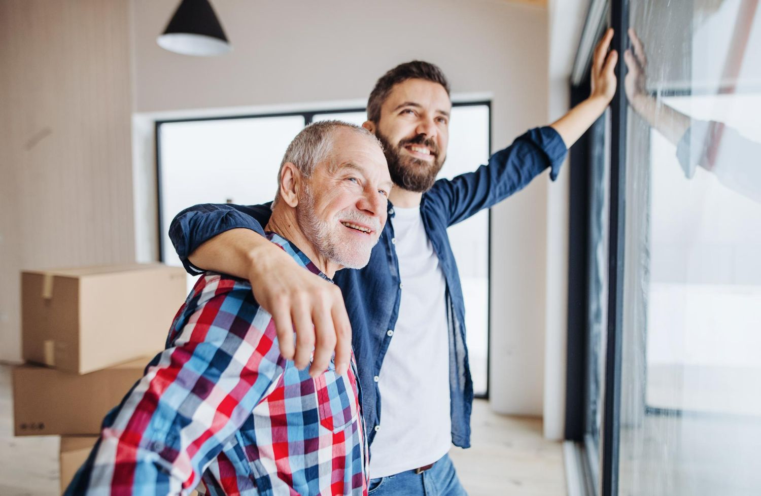 Two smiling people in a bright apartment, one with an arm around the other near moving boxes.