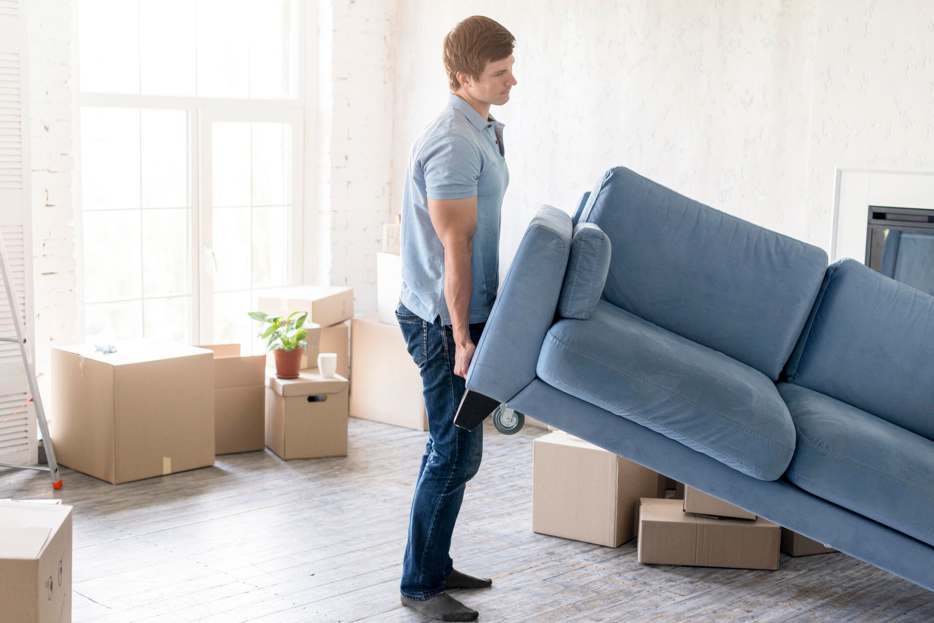 Man lifting a blue sofa in a room with cardboard boxes.
