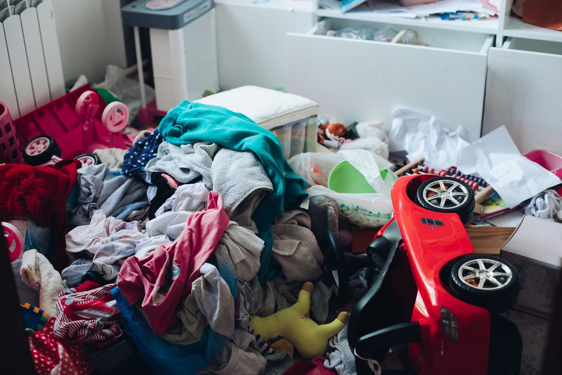 Pile of clothes and toys scattered on the floor in a messy room. A red toy car is visible.