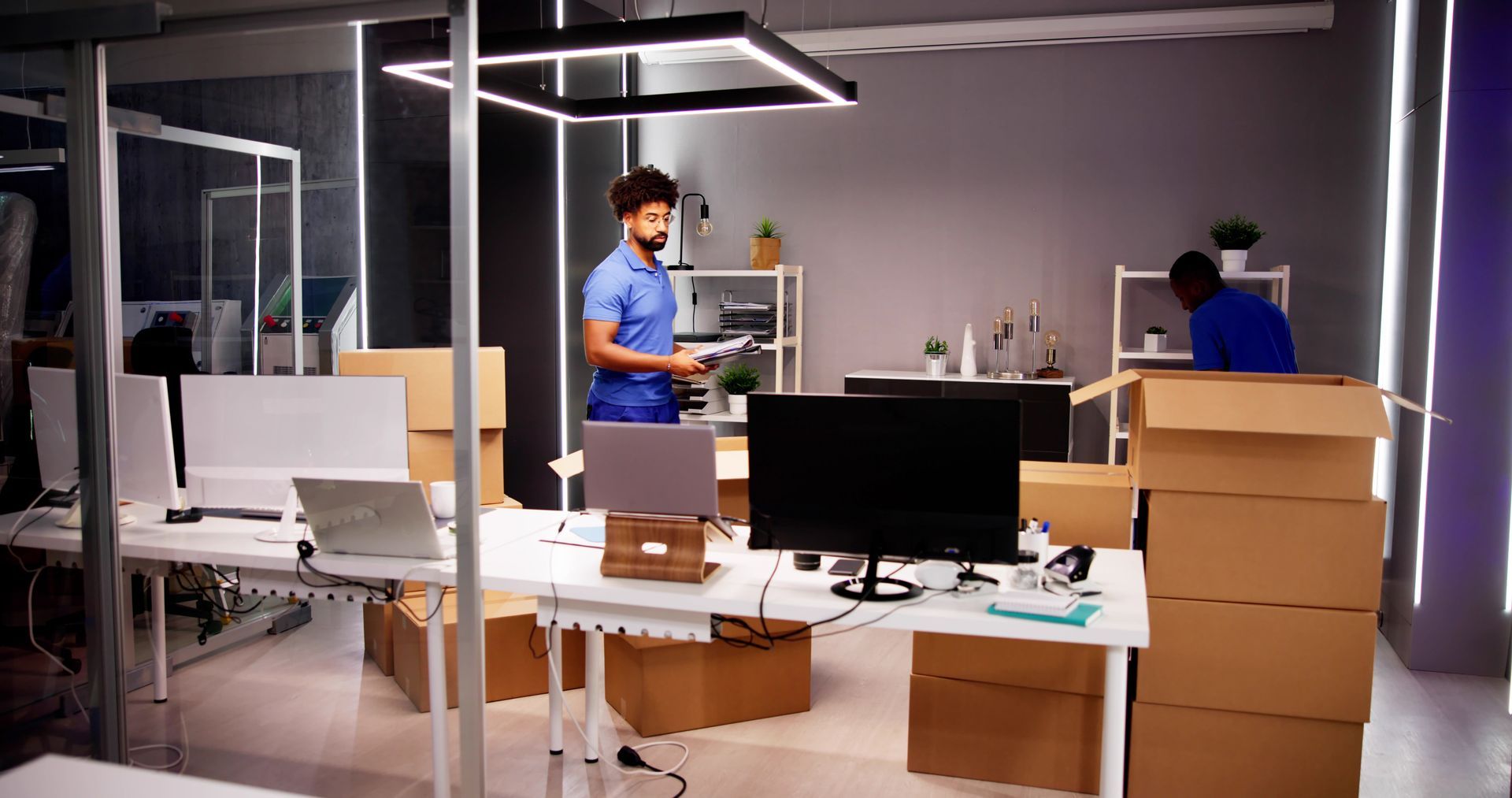 Two people in office, unpacking boxes. Desks and computers are visible. Fluorescent lights.