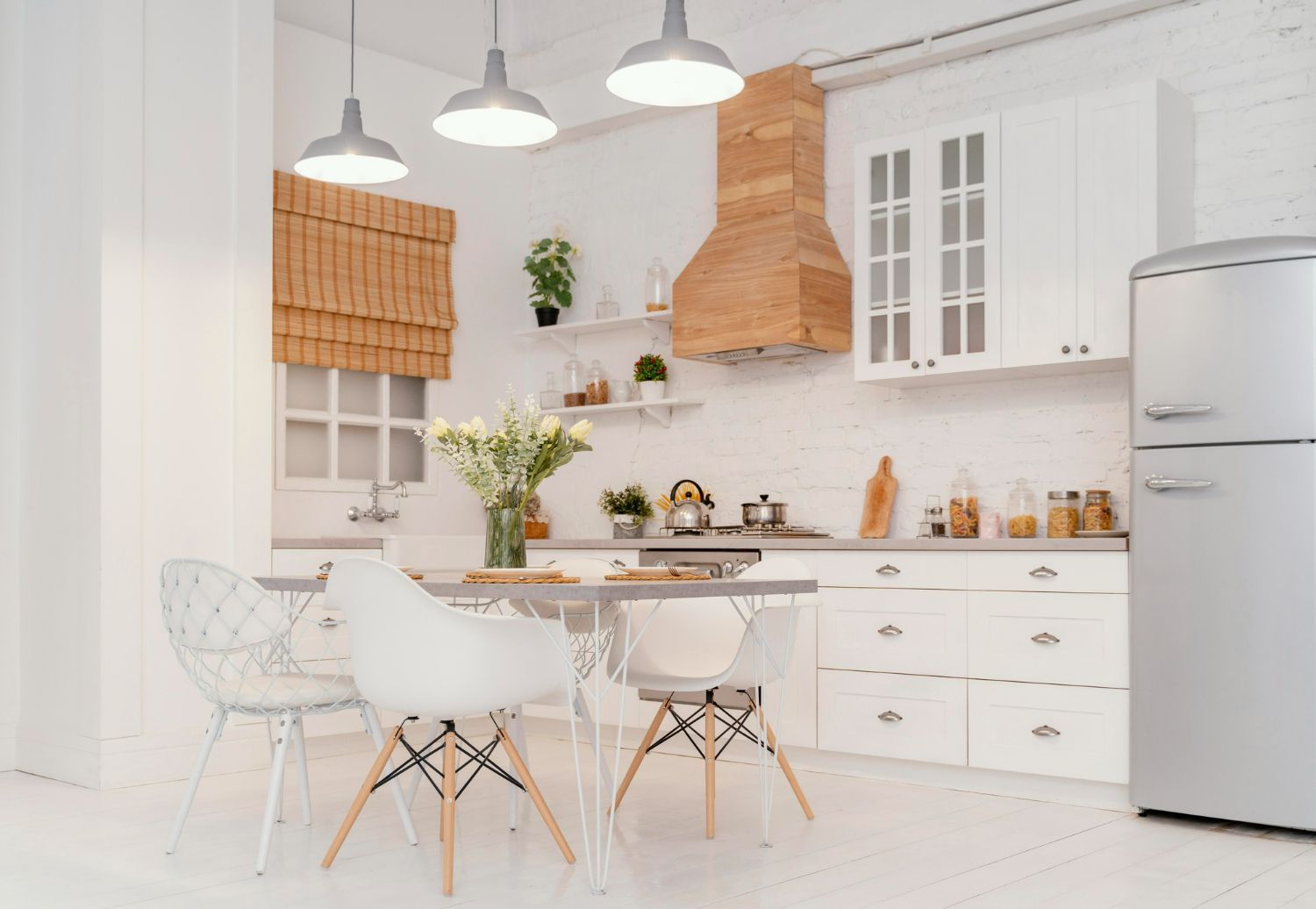 Bright, white kitchen with a dining table, chairs, and refrigerator. Wooden accents and hanging lights.