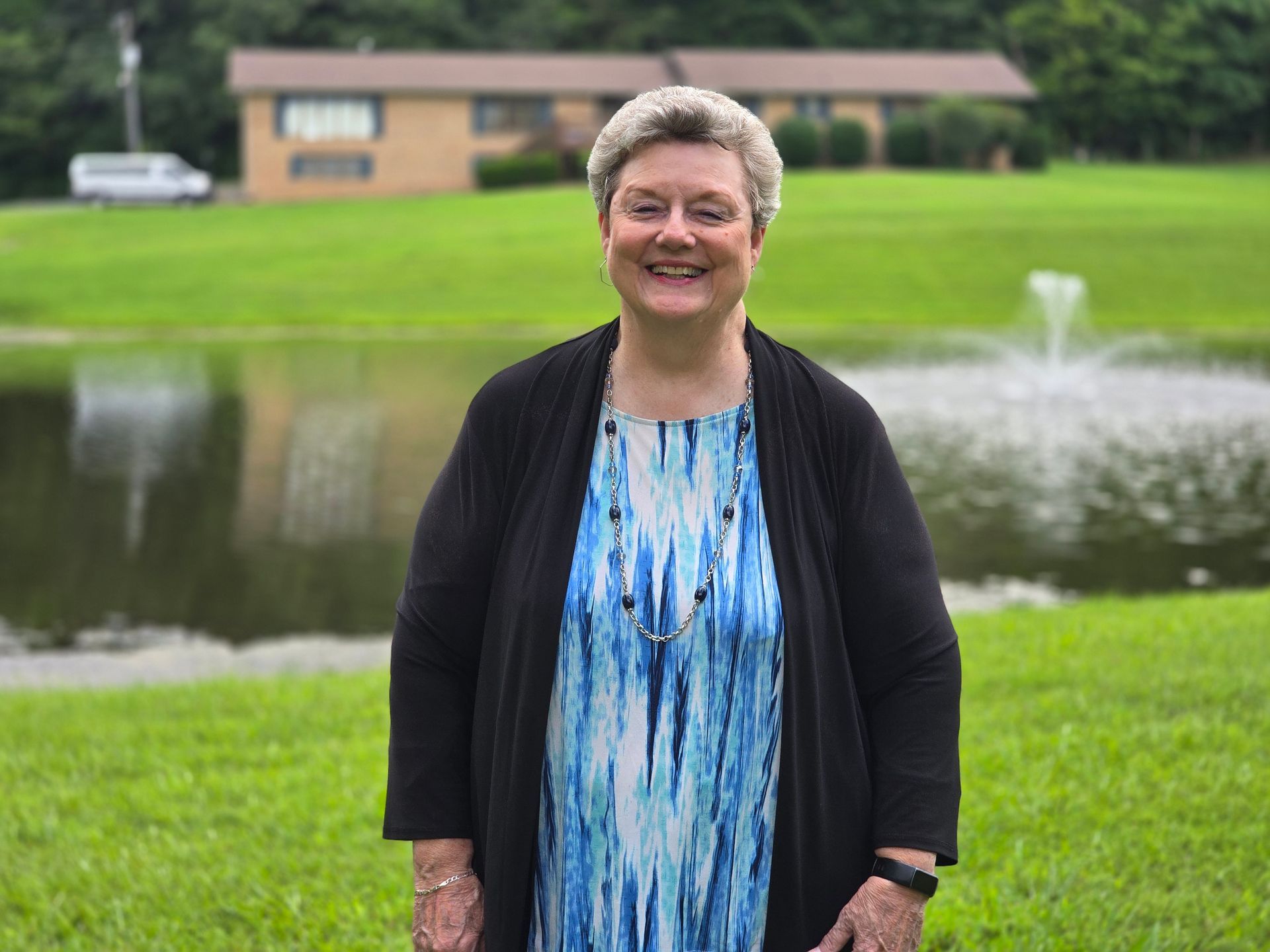 Smiling person in a blue dress and black cardigan standing by a pond with a fountain and building in the background