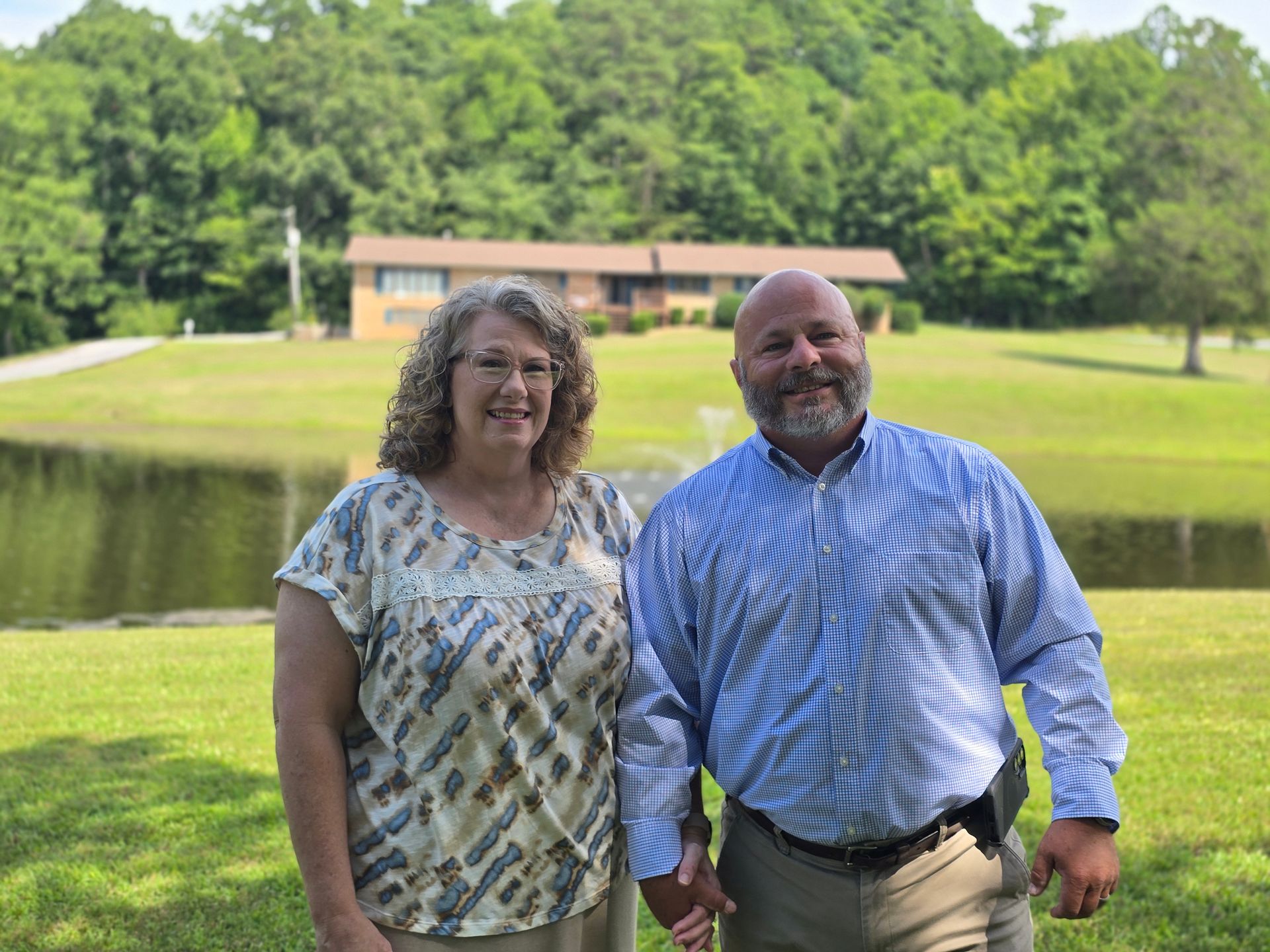 Two people smiling by a pond, with a building and green trees in the background.