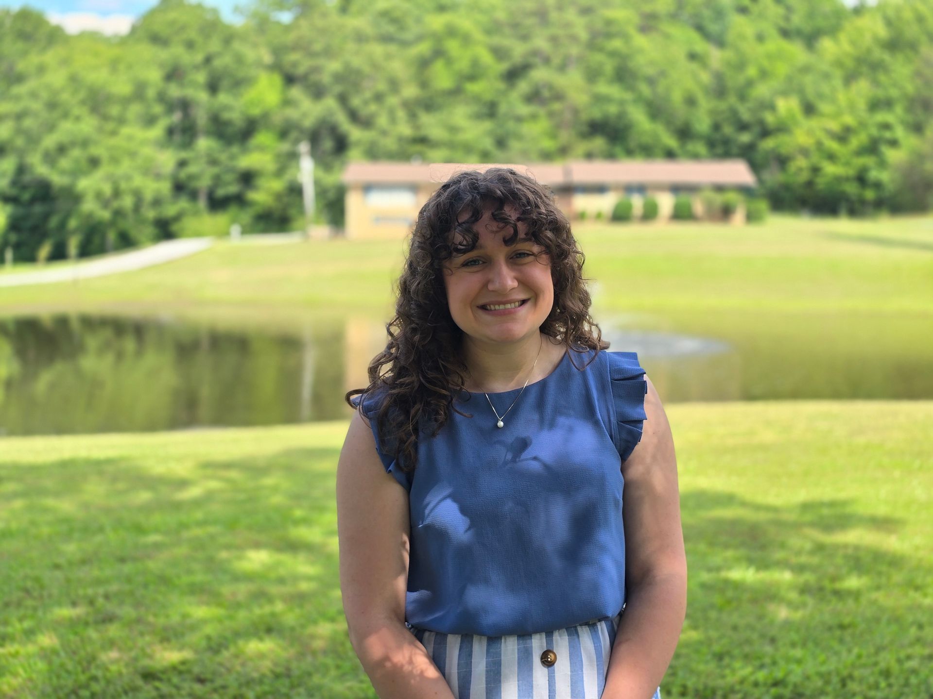 Woman smiling in a blue dress standing on grass near a pond with trees and a building in the background