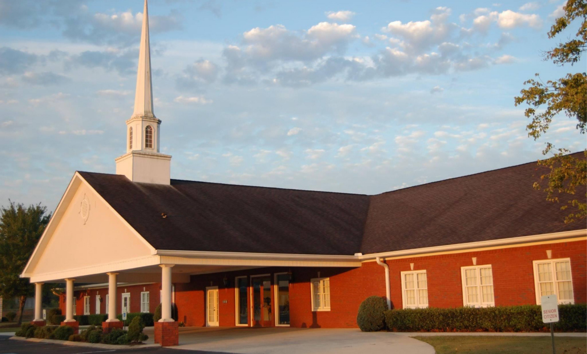 White church with steeple, red brick walls, and brown roof under a cloudy sky