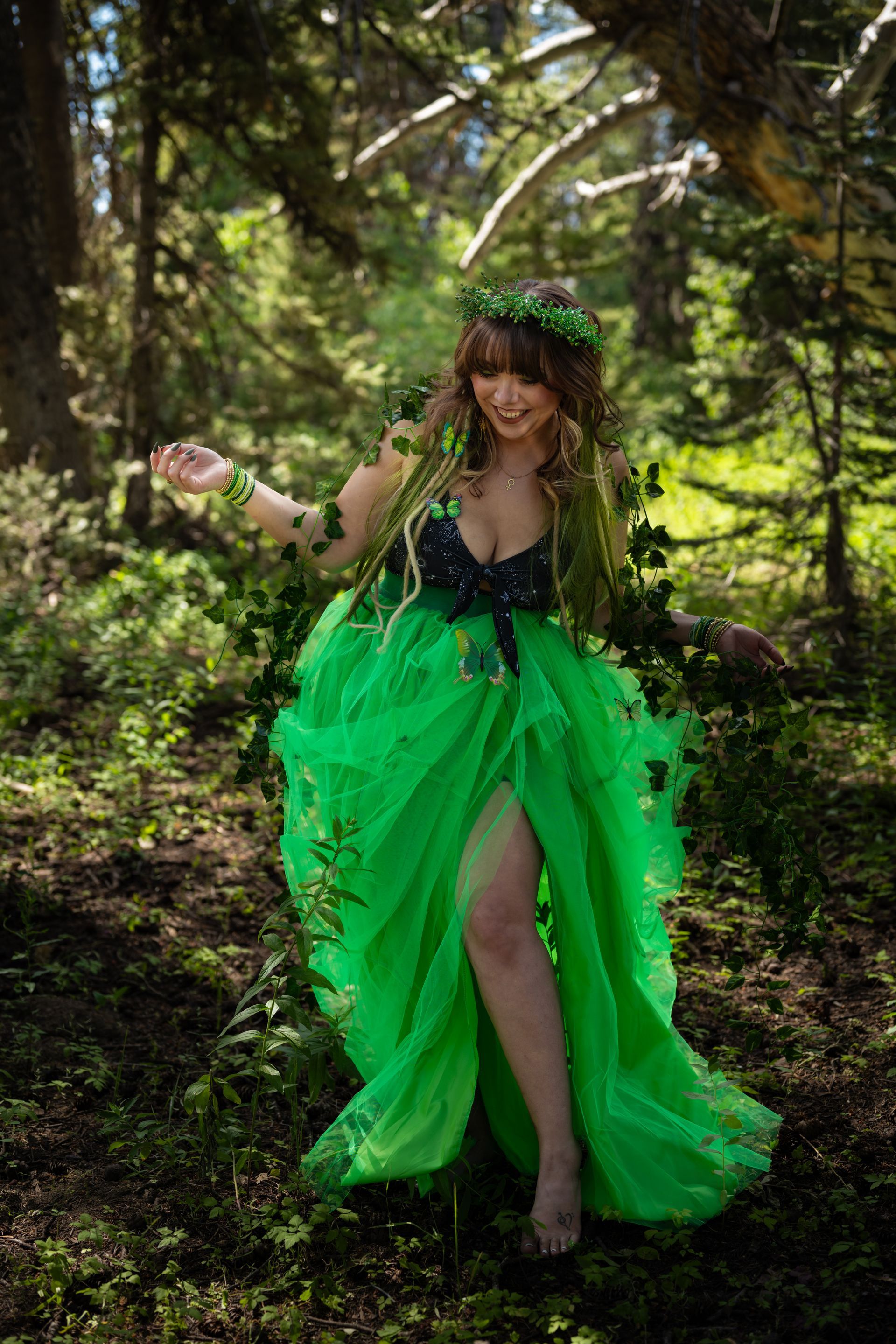 A witch in a bright green dress posing in a sunlit forest clearing with arms outstretched.