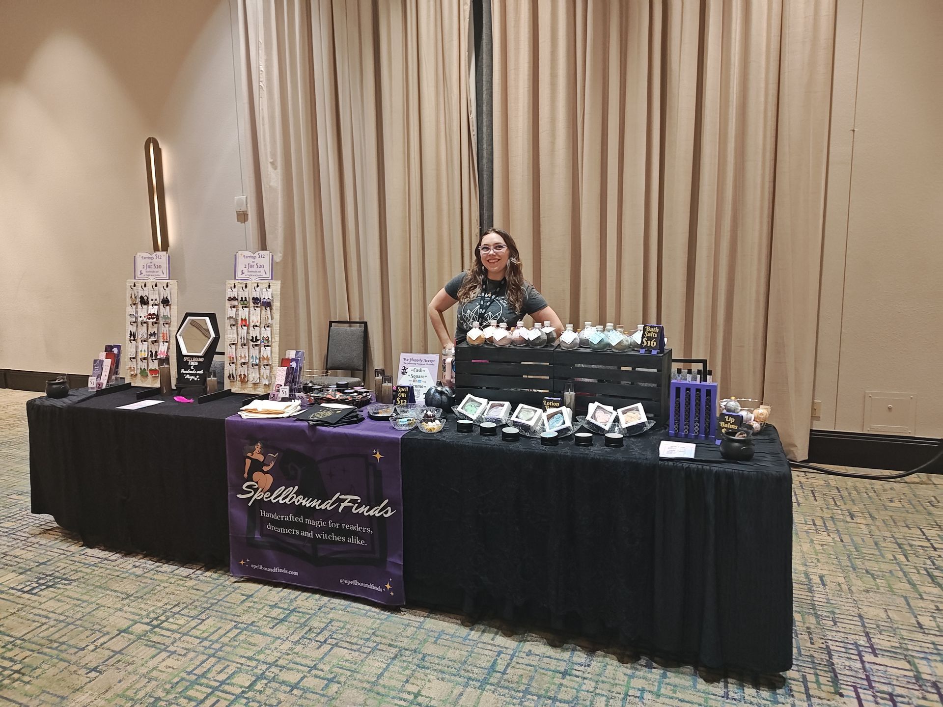 Vendor booth with black tablecloth, display items, and a person standing behind it in a beige conference hall