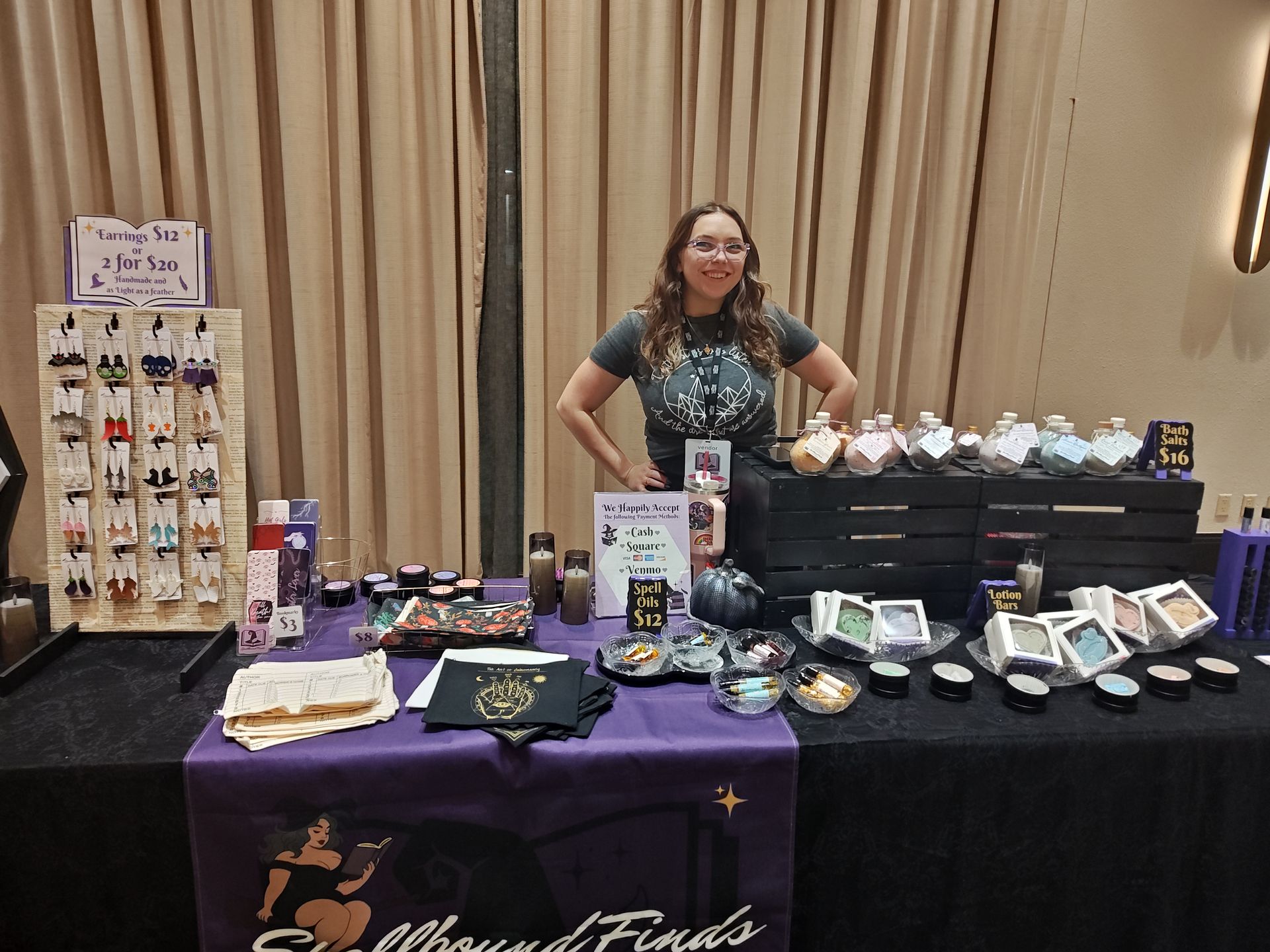 Vendor booth with crystals and jewelry displayed on tables, woman standing behind it in a curtained room
