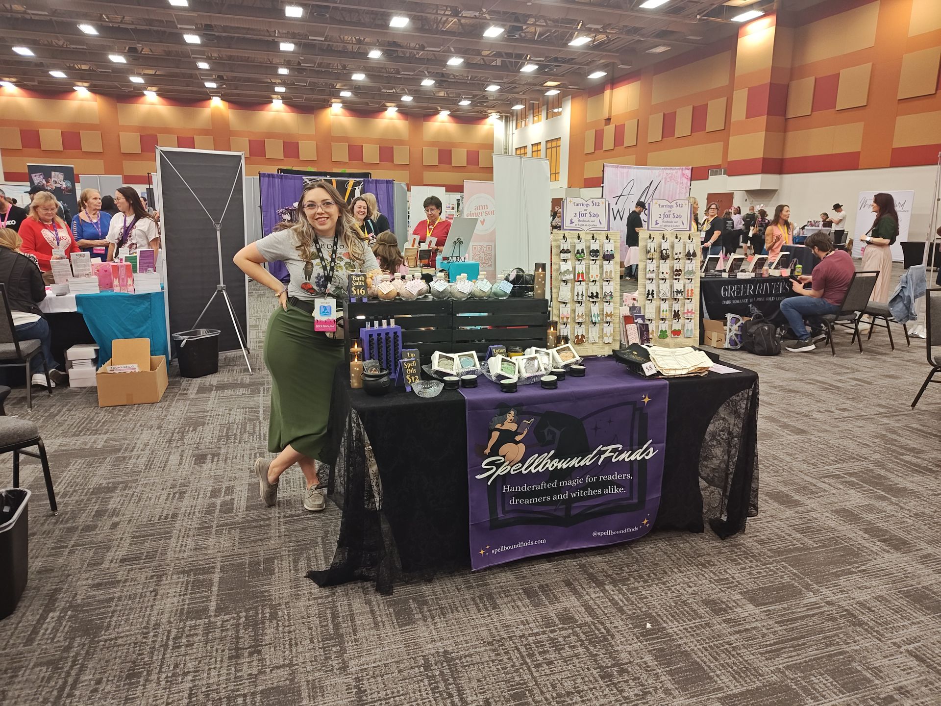 Vendor booth at a craft fair, with jewelry display and a woman posing beside the table