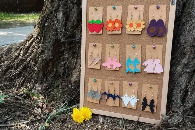 Handmade earrings displayed on a cork board outdoors beside a tree and yellow flowers.