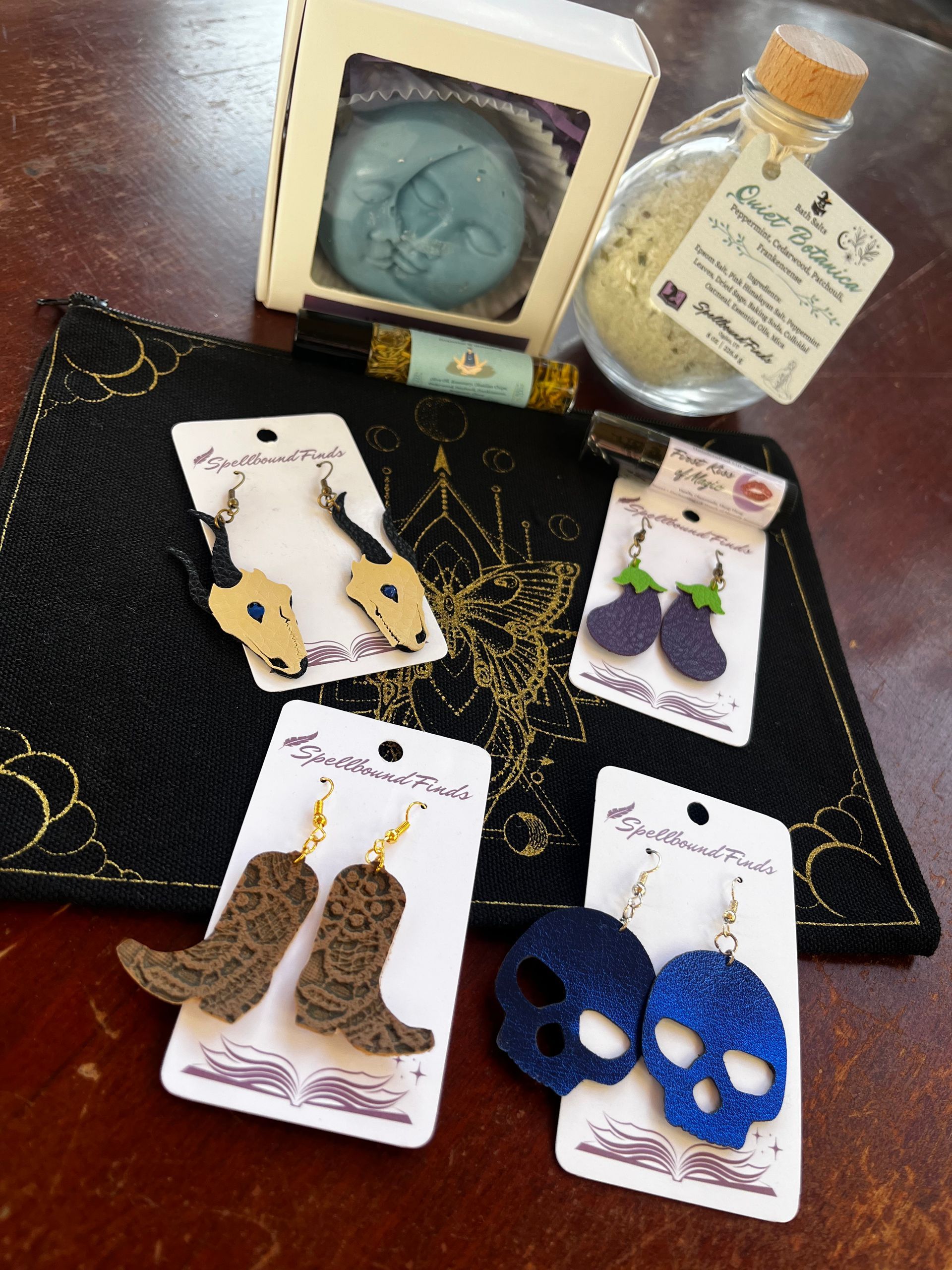 Assorted handmade earrings on cards, including boots, skulls, and cats, arranged on a dark cloth with a candle and jar.