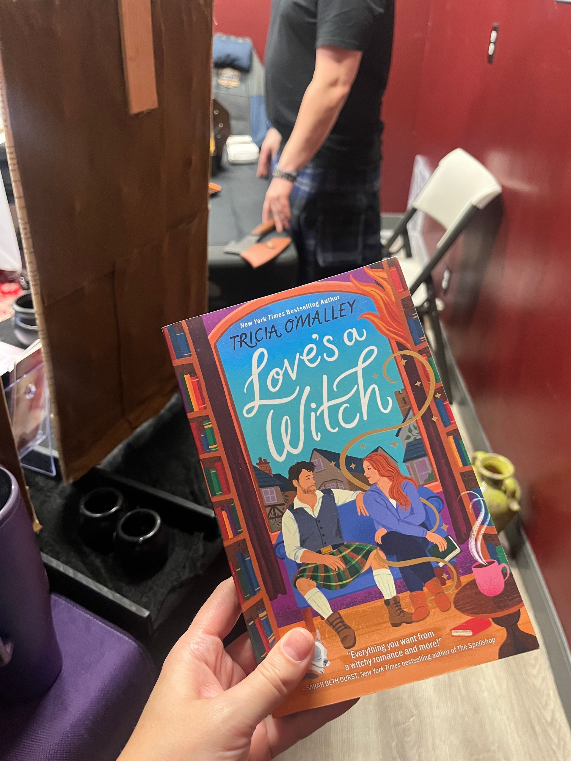 Hand holding the book “Love’s a Witch” in front of a convention booth backdrop