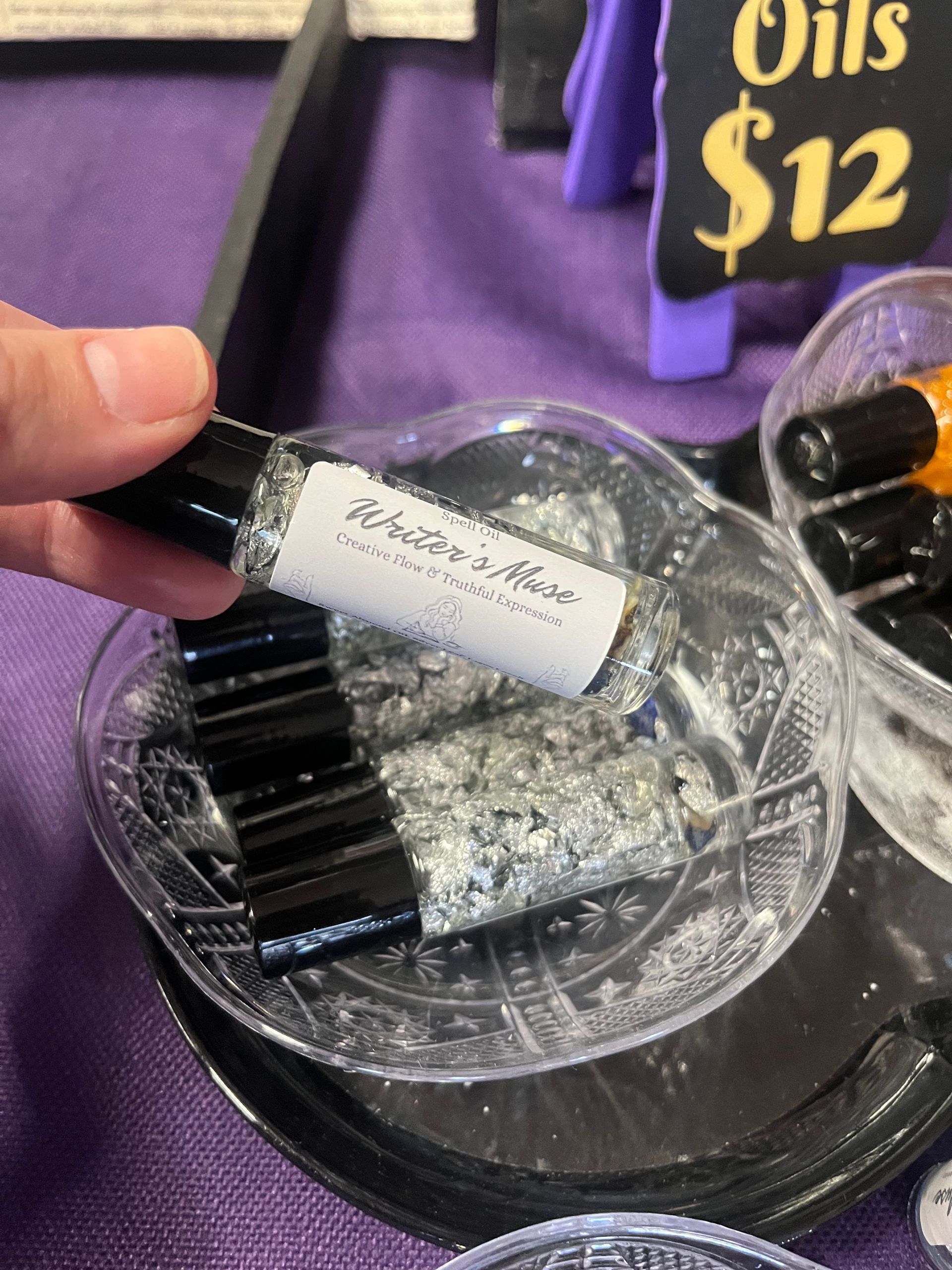 Hand holding a lip gloss tube over a display of silver glitter nail polish bottles beside a “$12” sign