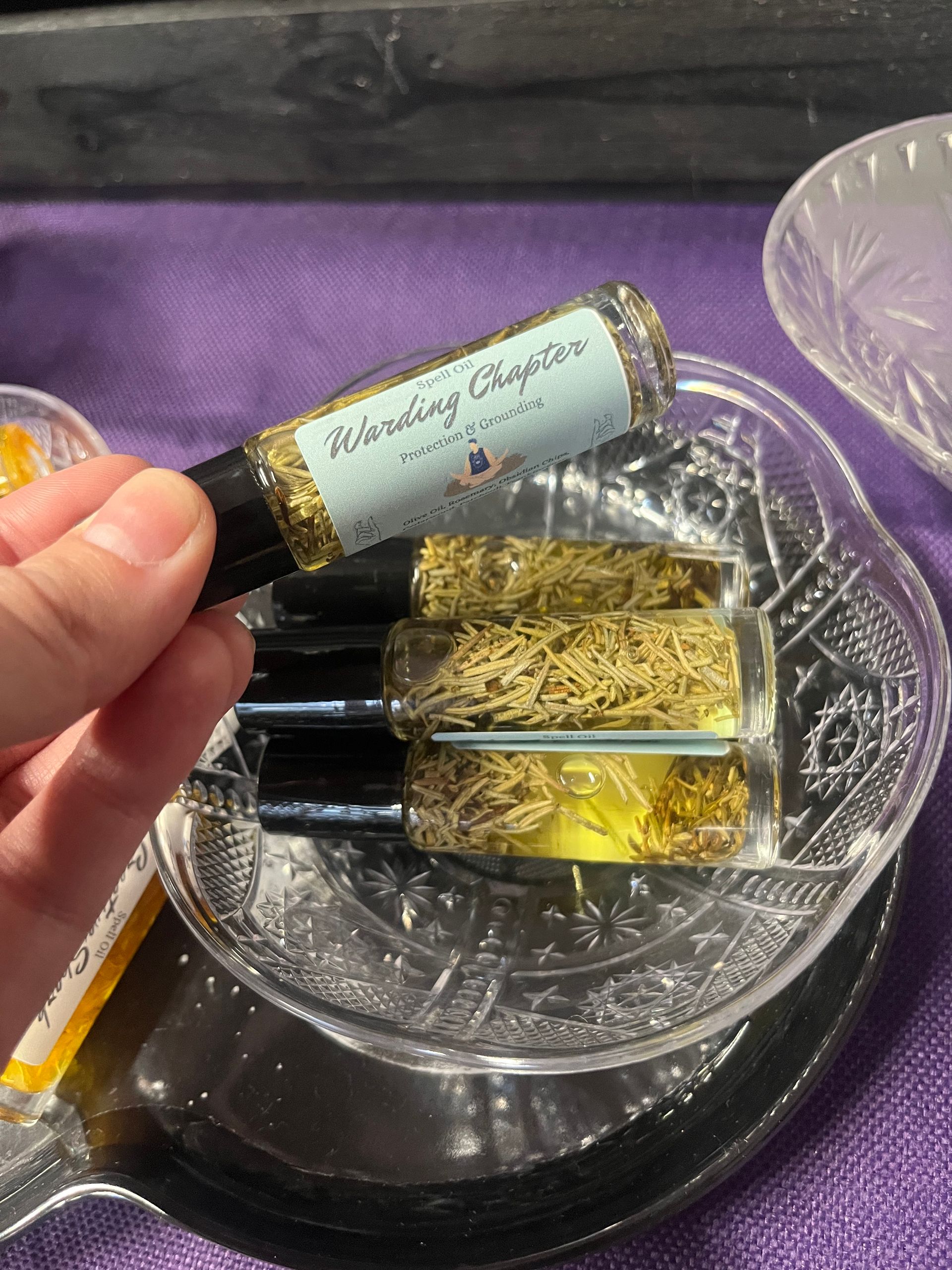 Hand holding two small glass vials over a crystal bowl on a purple cloth, with a gold sparkly liquid inside.