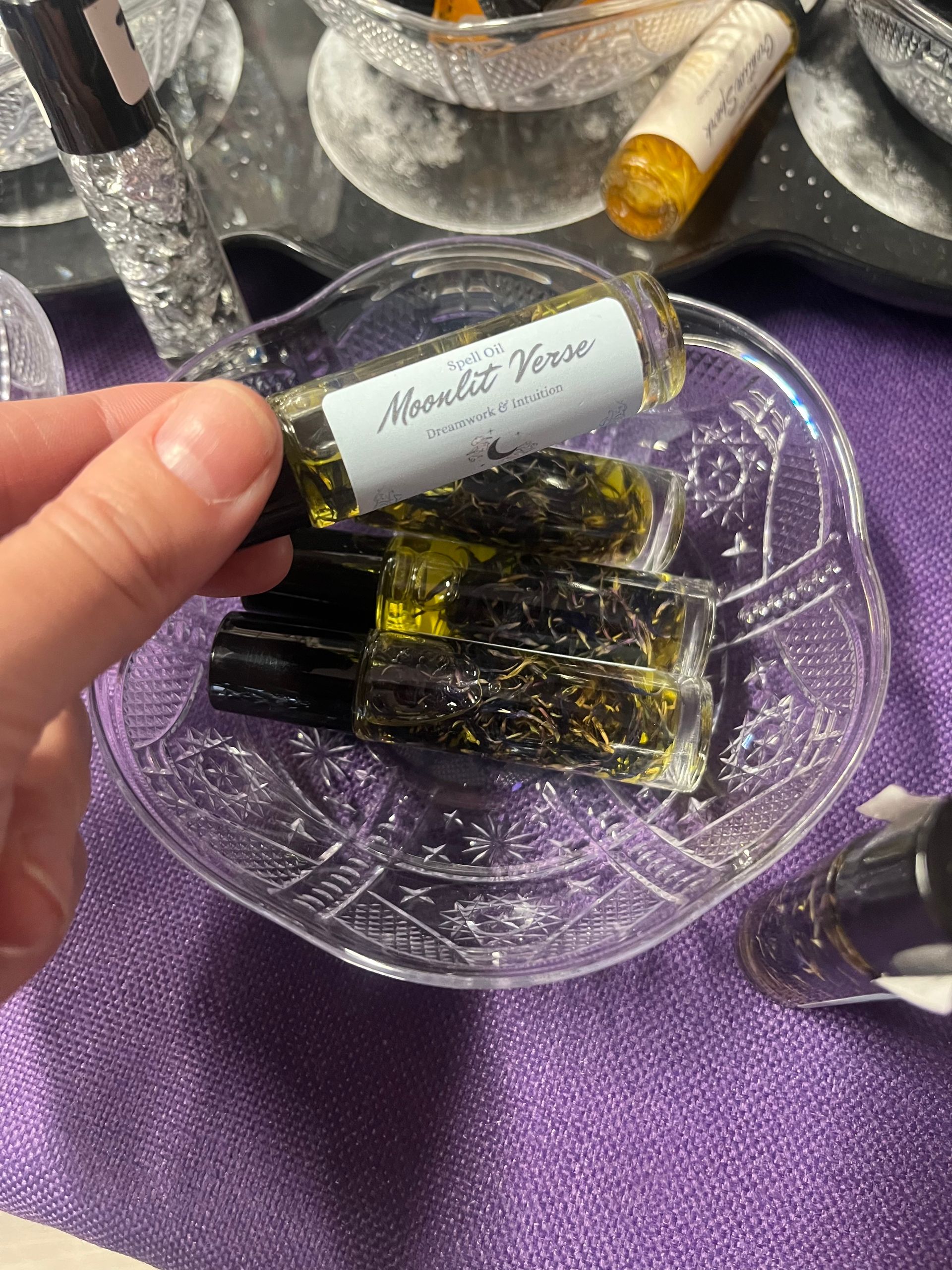 Hand holding a small glass perfume vial above a purple cloth with several black-and-gold vials below