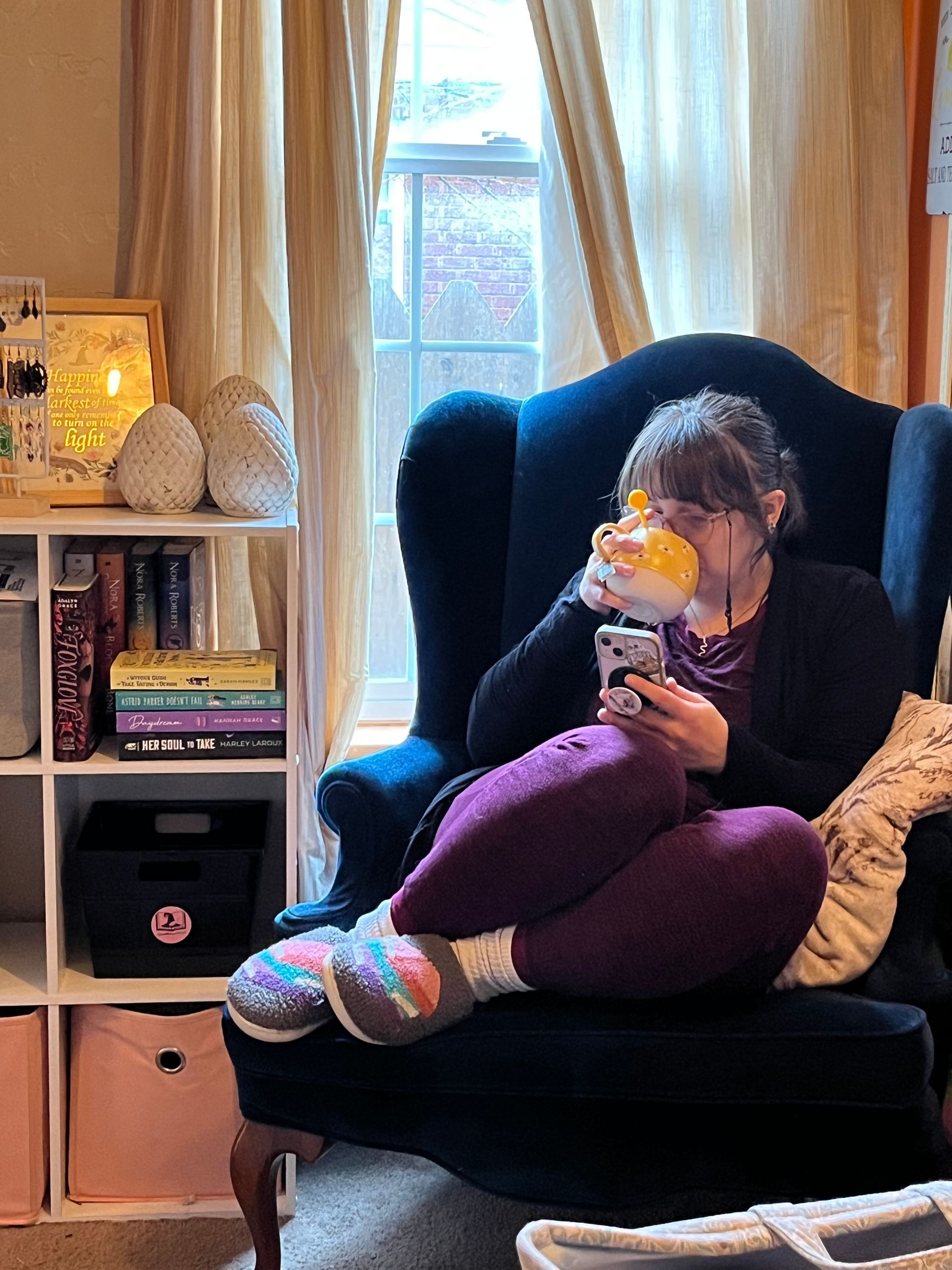 Person sitting in a dark blue armchair by a window, holding a stuffed toy, with shelves and a lamp nearby.
