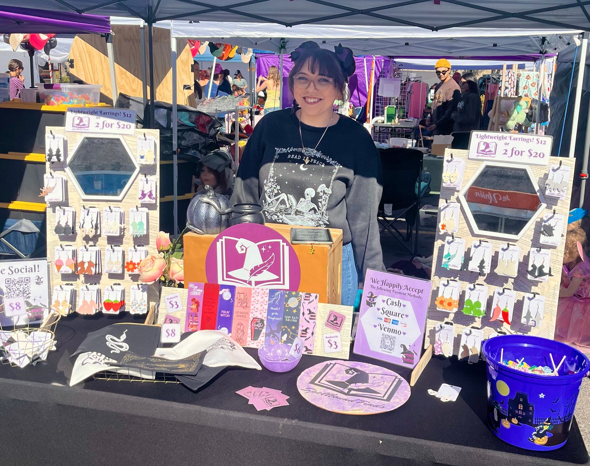 Vendor smiling behind craft booth with colorful handmade items under a market tent