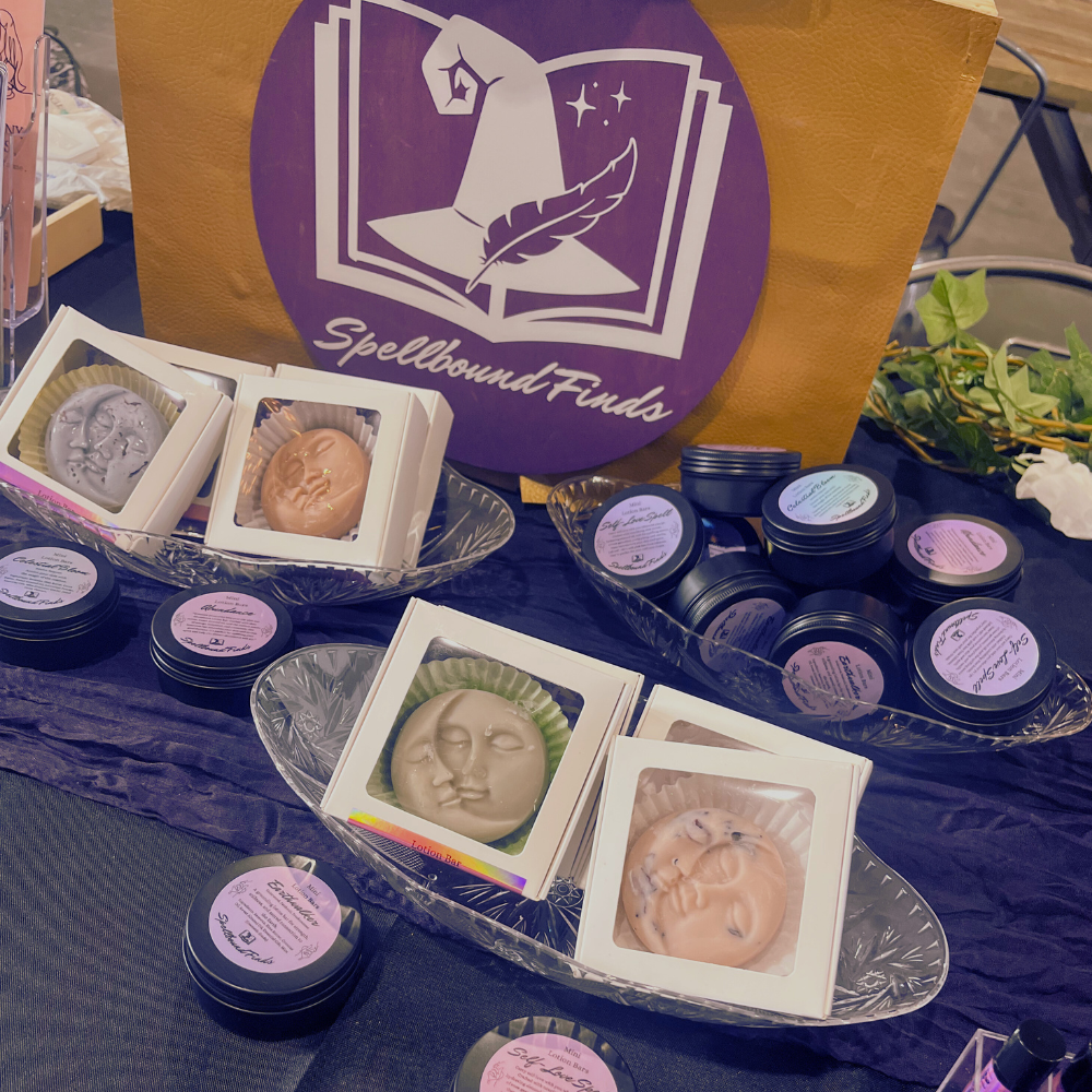 Assorted handmade soaps and bath products displayed in trays with a purple logo bag behind them