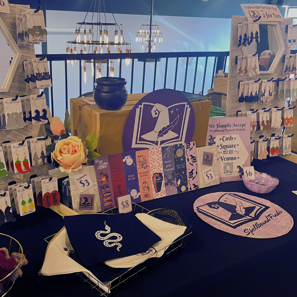 Book-themed vendor display with black-and-purple stationery, tote bags, candles, and decorative shelves