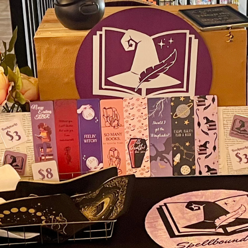 Purple and black vendor display of spellbook-themed bookmarks, cards, and trays on a table
