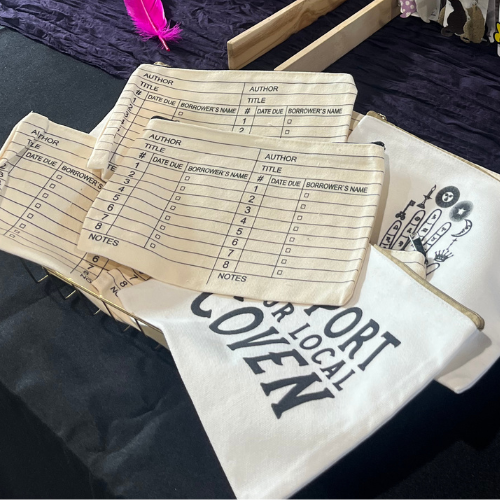 Printed tickets and a white tote bag with black text on a dark table, with a pink feather nearby