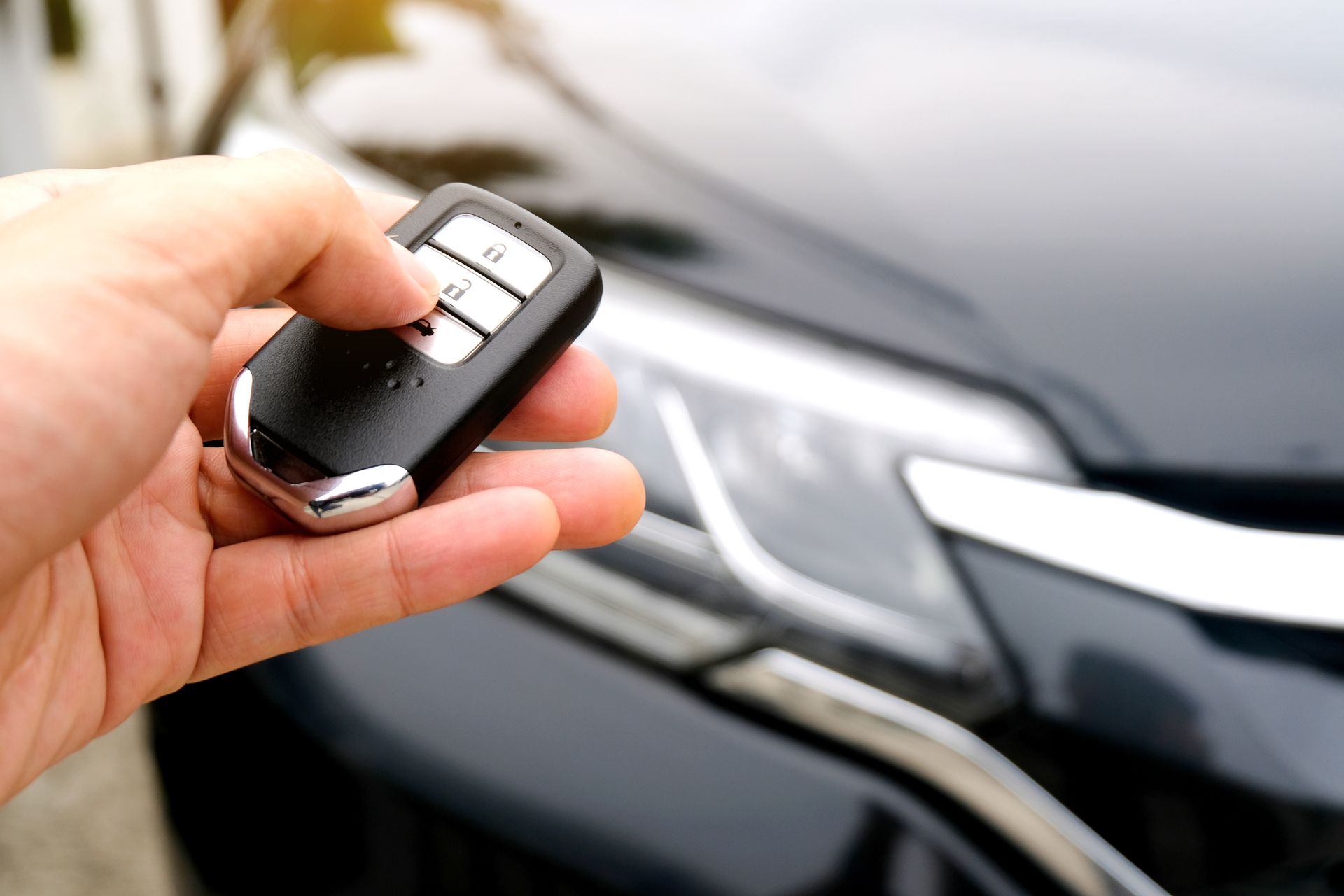 Hand holding a car key fob near a black vehicle’s front grille. Hand holding a car key fob near a black vehicle’s front grille.