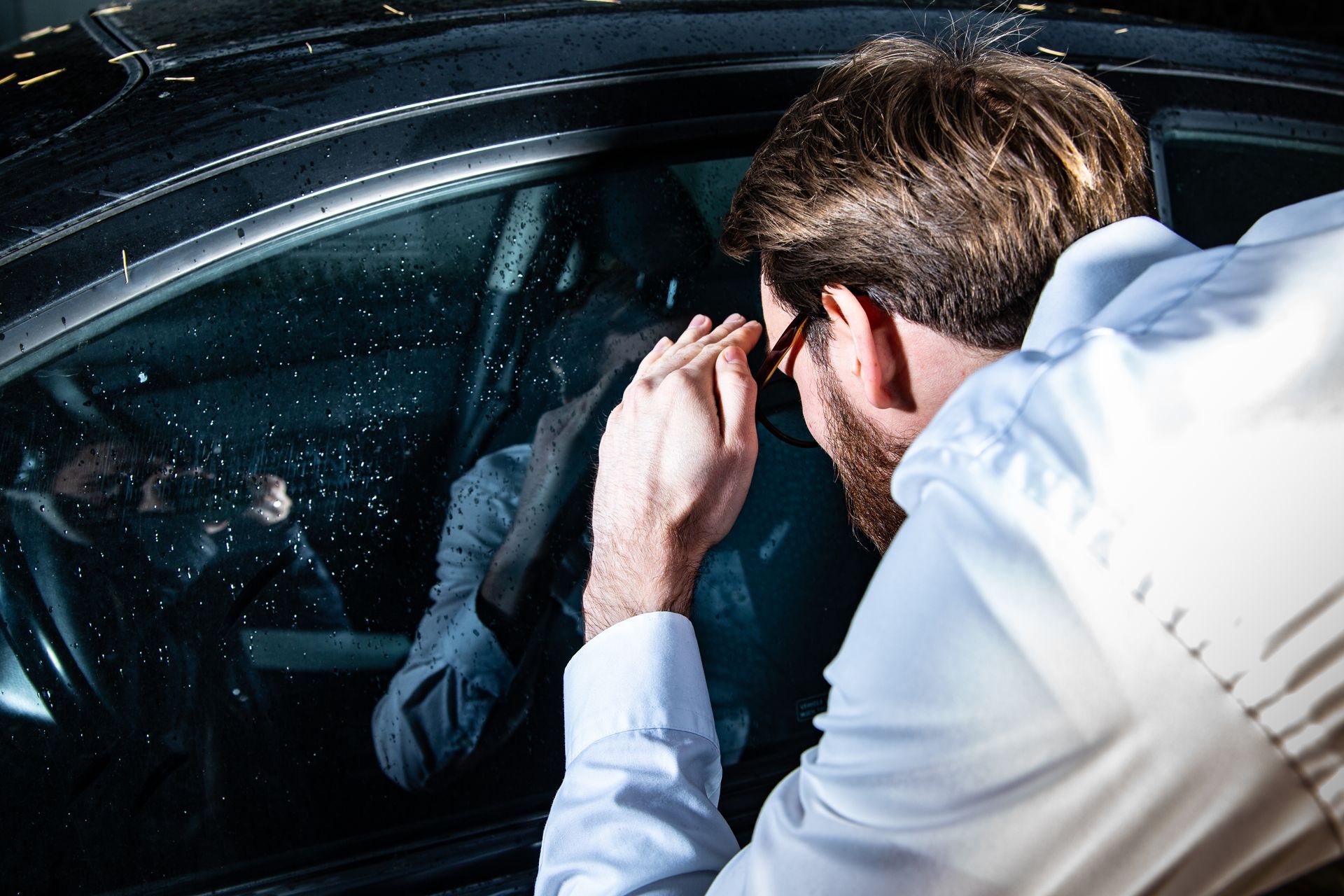 Man in a white shirt peers into a car window, adjusting glasses beside a shadowed person inside. Man in a white shirt peers into a car window, adjusting glasses beside a shadowed person inside.