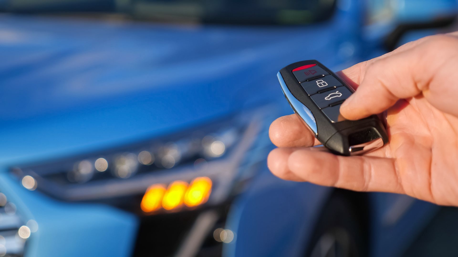 Hand holding a car key fob in front of a blue car with headlights on. Hand holding a car key fob in front of a blue car with headlights on.