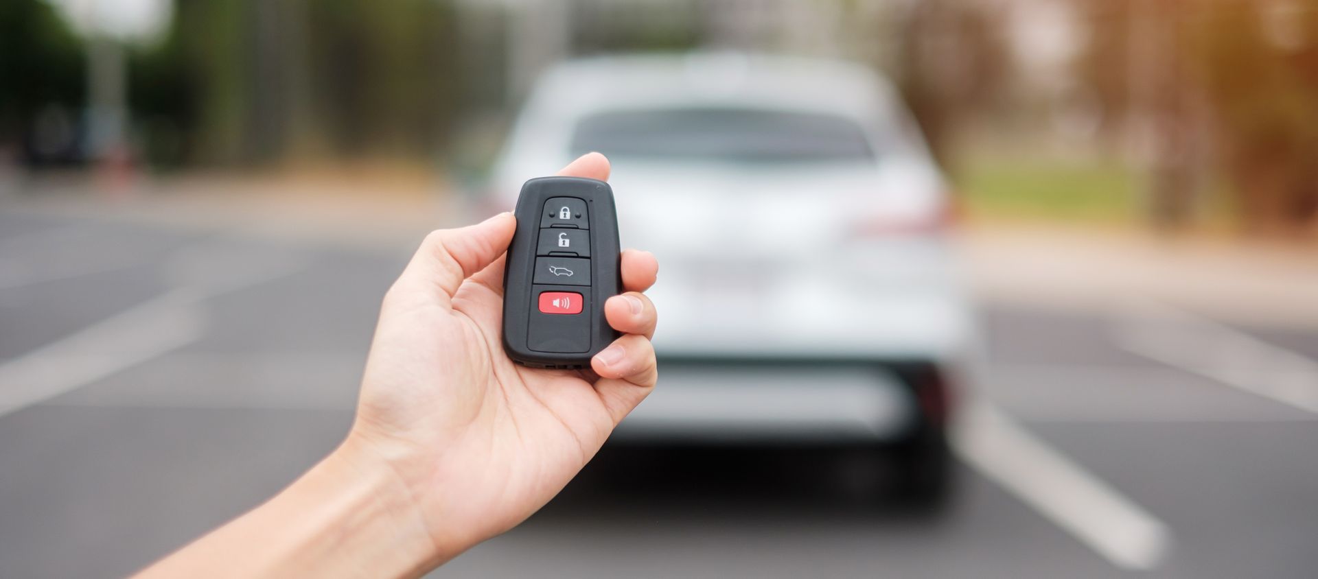 Hand holding a car key fob toward a white car in a parking lot. Hand holding a car key fob toward a white car in a parking lot.