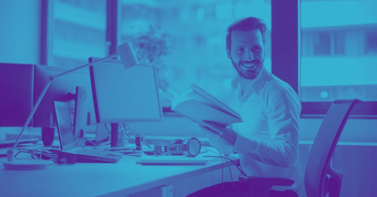 A man is sitting at a desk in front of a computer holding a book.