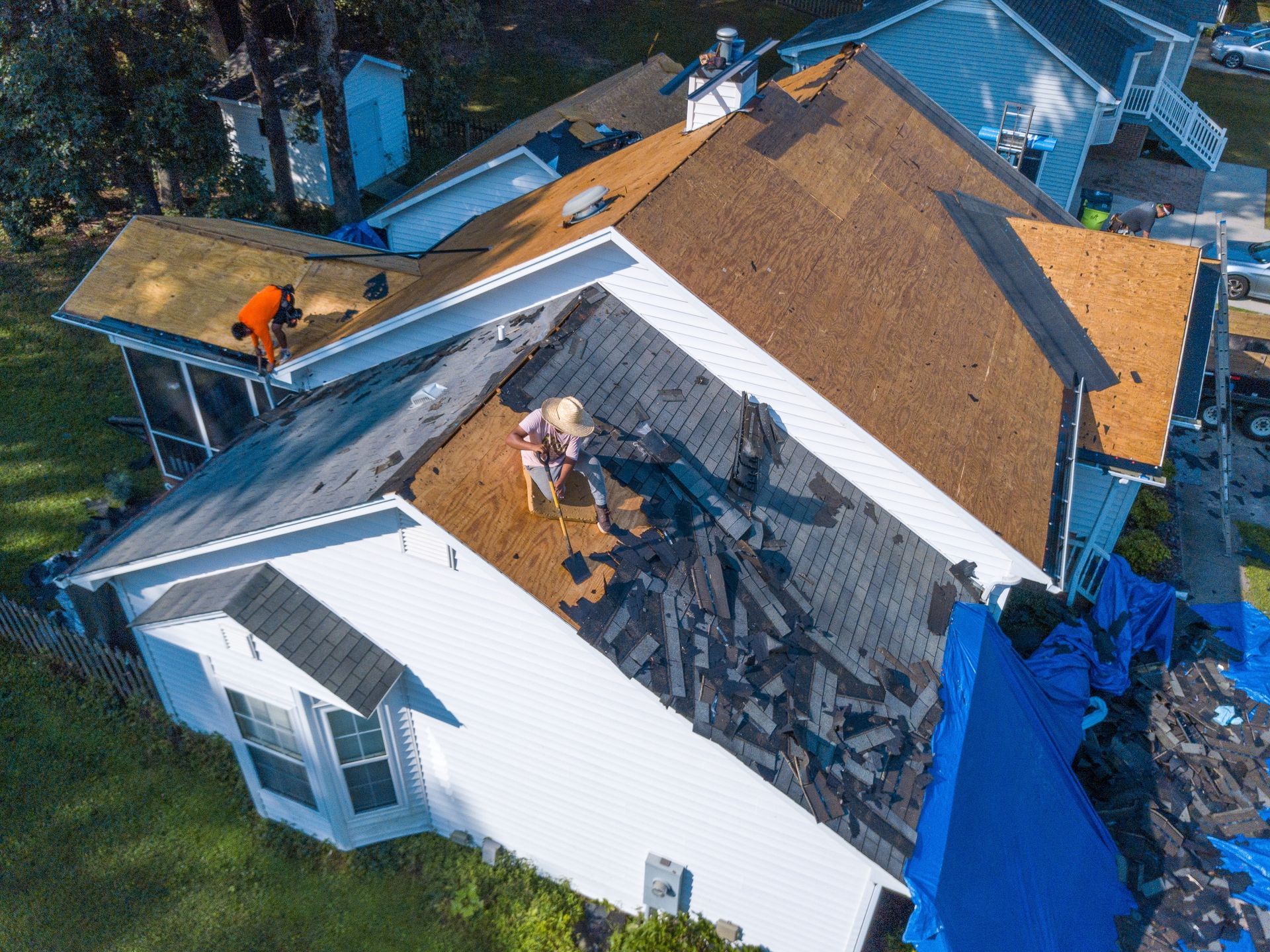 Workers replacing a roof on a white house; one on the left, two in the middle. Blue tarp covers part of the roof.
