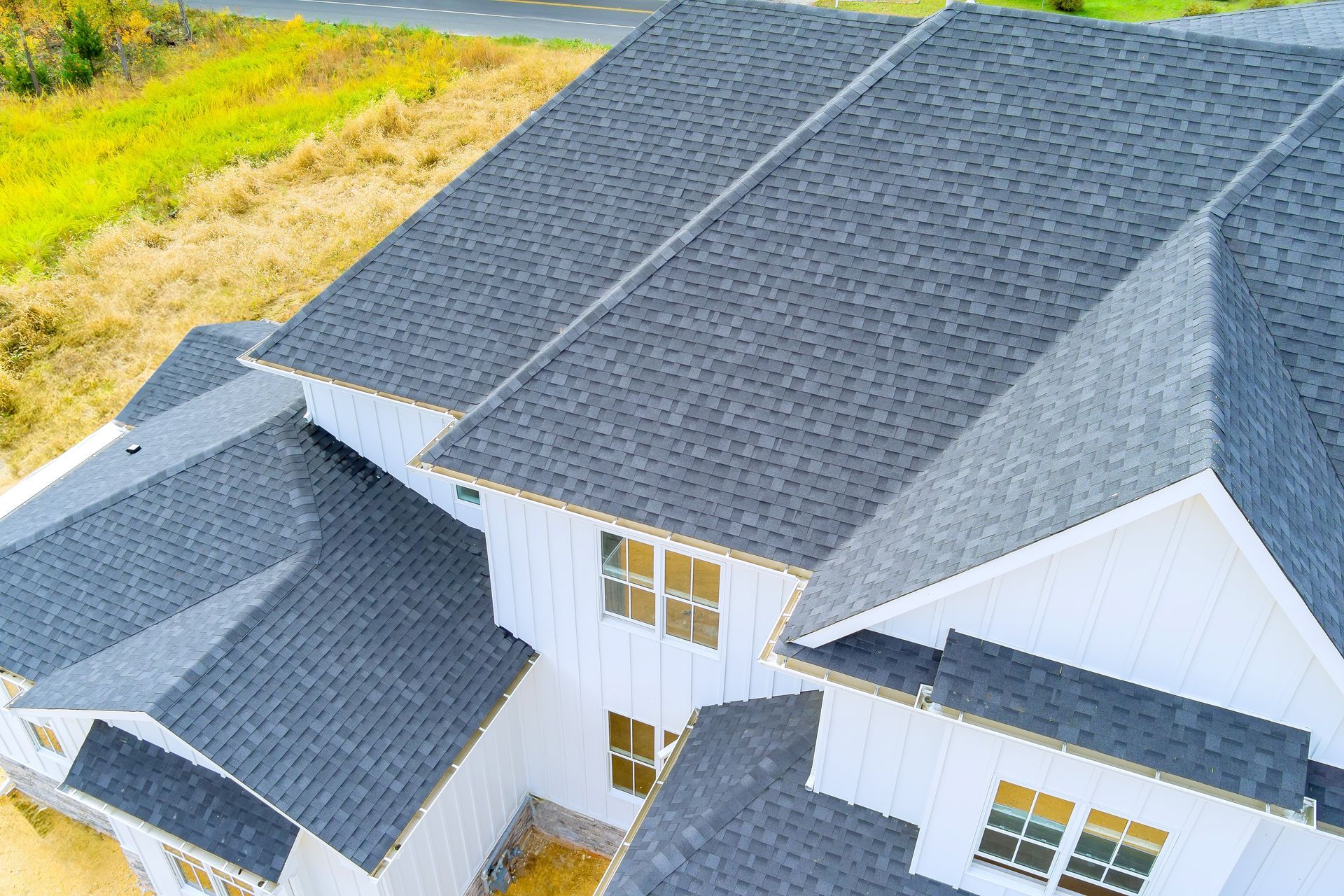 Aerial view of a white house with a dark gray shingled roof, windows, and surrounding green field.