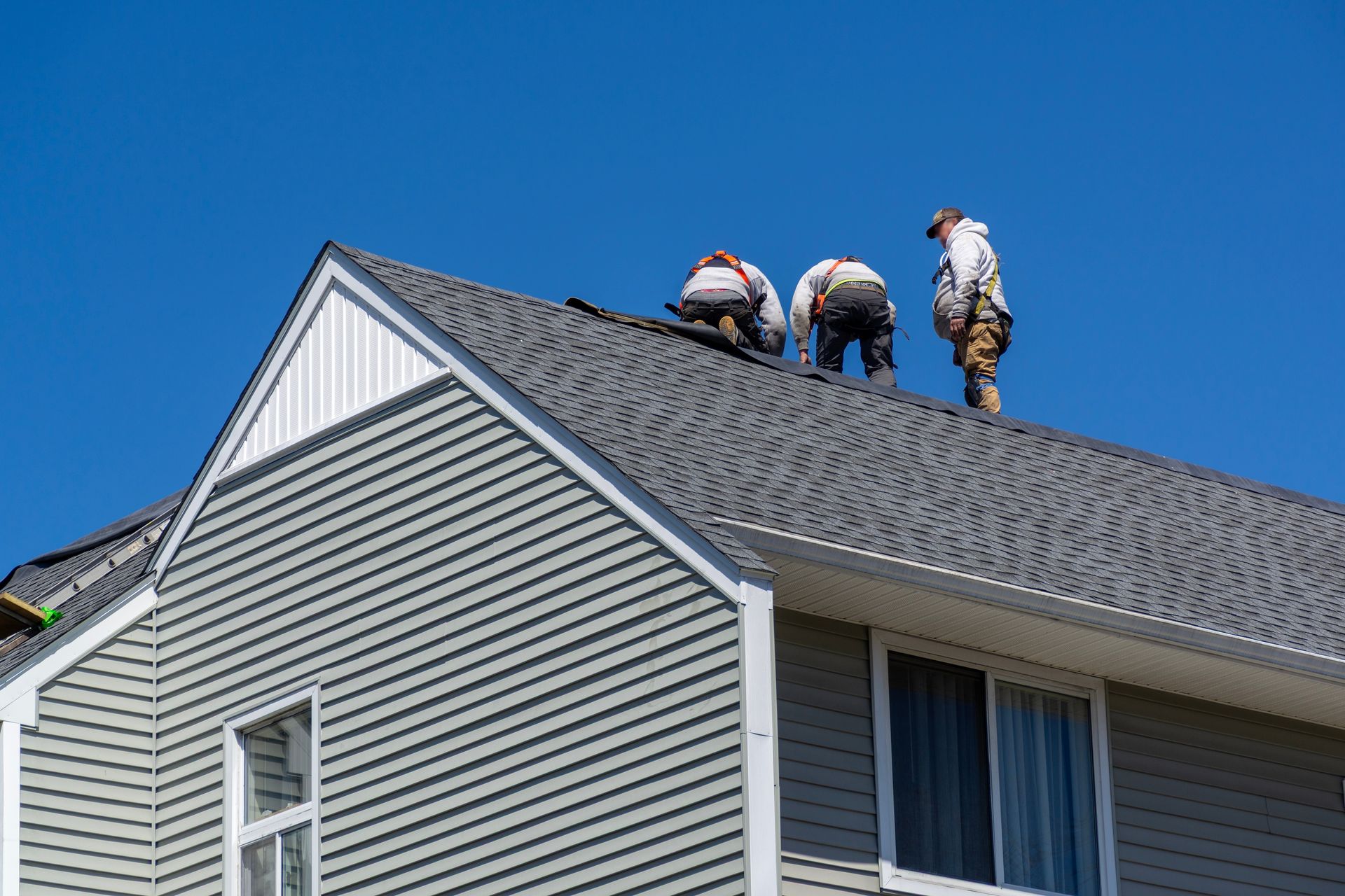 Three roofers on a gray shingled roof, against a blue sky. They wear safety harnesses.