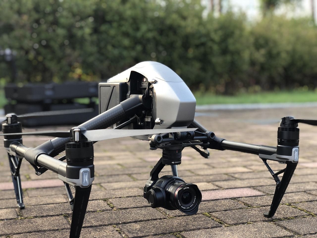 Drone on pavement with camera, black arms, gray body, green background.