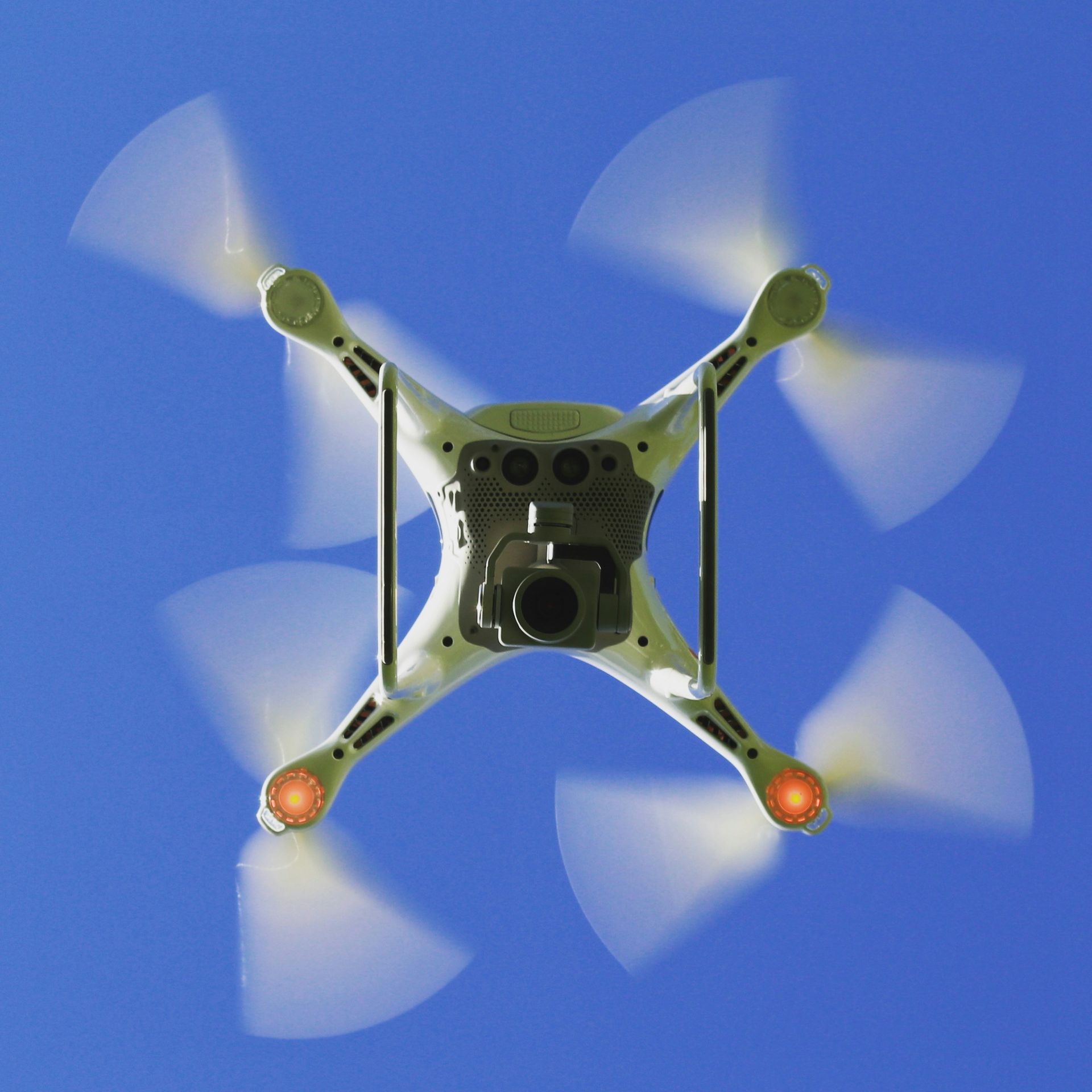 Drone with spinning blades, camera facing down, against a clear blue sky.