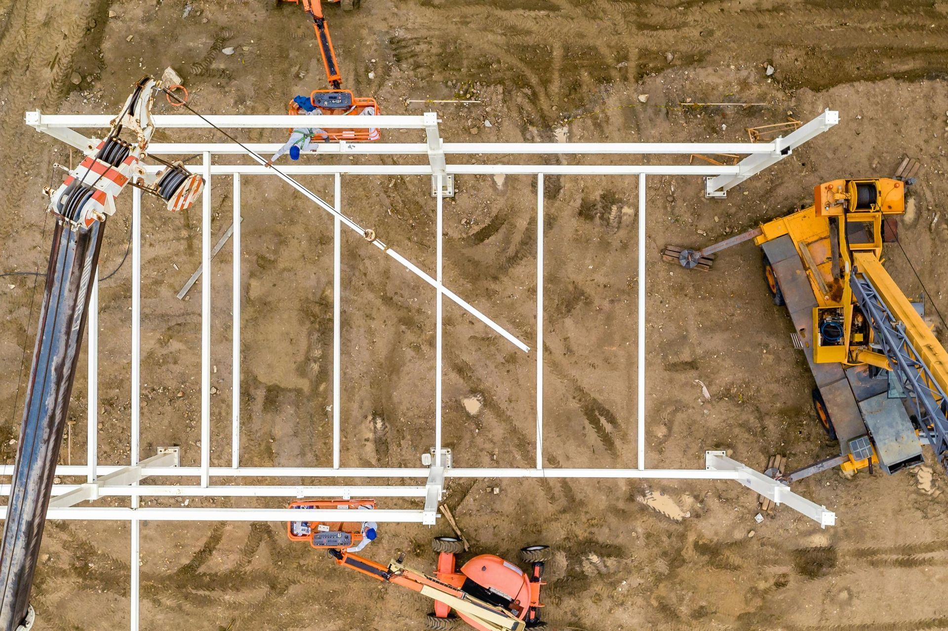Overhead view of construction site with white metal framework being assembled by cranes and machinery.