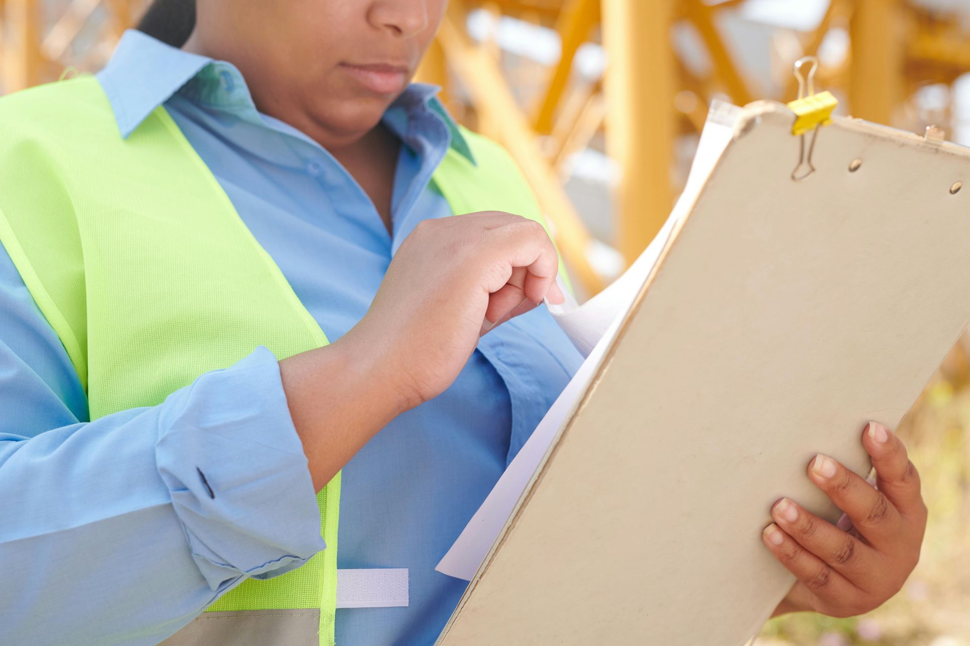 Woman in safety vest examines papers on a clipboard, construction site background.