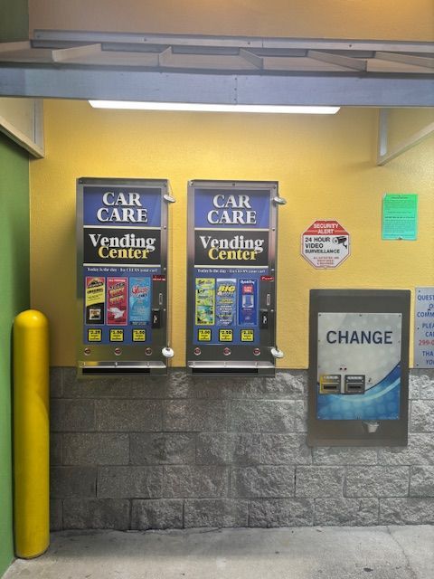 Two car care vending machines and a change machine on a yellow and stone wall.