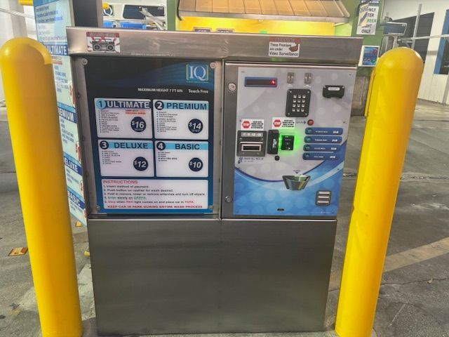 Car wash payment machine with yellow bollards. Offers four wash packages: Ultimate, Premium, Deluxe, and Basic.