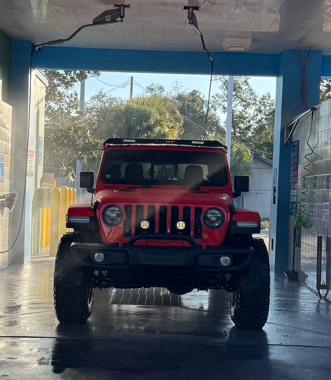Red Jeep Wrangler at a car wash, water spraying, under a blue canopy.