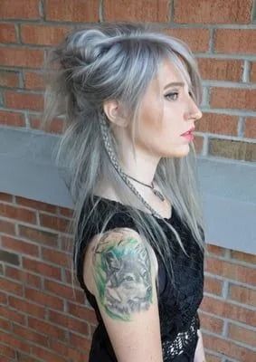 Profile of a person with silver hair and tattoos, standing beside a brick wall indoors.