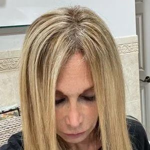 Person with long blonde hair looking down in a bathroom mirror, wearing a black top