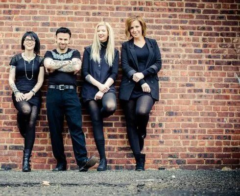 Four people posing against a brick wall, dressed in dark clothing, with relaxed expressions.