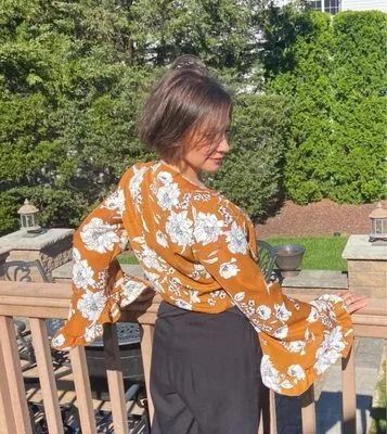 Person in an orange floral blouse leaning on a wooden railing in a sunny garden.