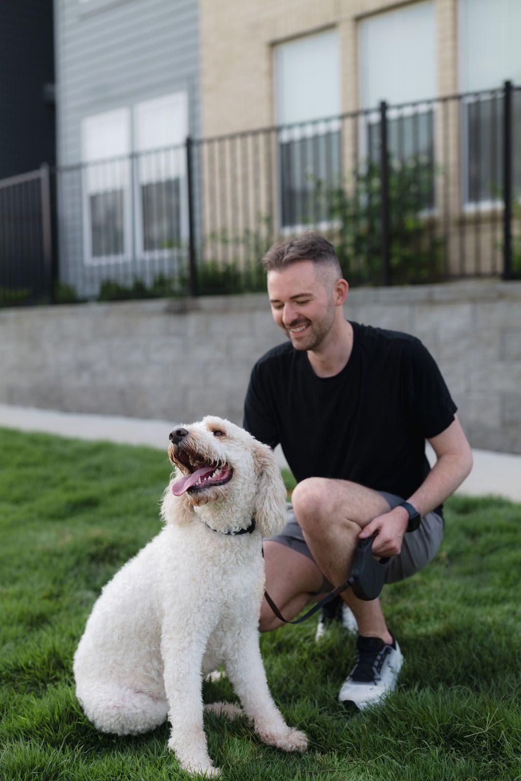 Man crouches on grass smiling beside a white curly dog in front of a building