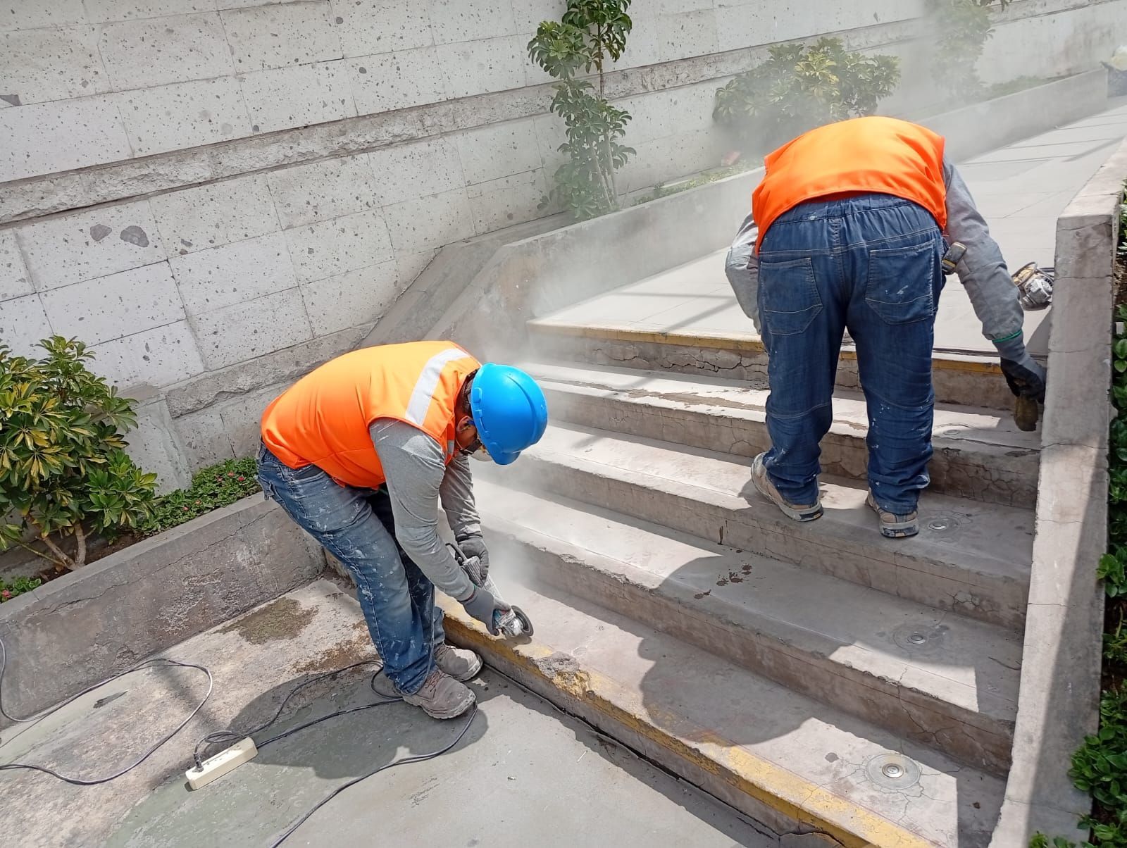 Obrero de la construcción con casco y chaleco de seguridad revisando planos junto a una base de hormigón y columnas de varillas de refuerzo.