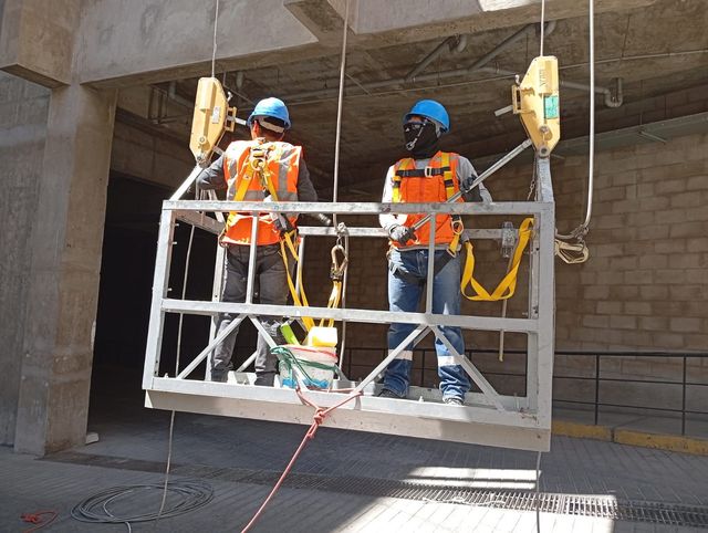 Dos trabajadores con chalecos de seguridad naranjas y cascos azules están de pie sobre una plataforma de trabajo suspendida en una obra de construcción.