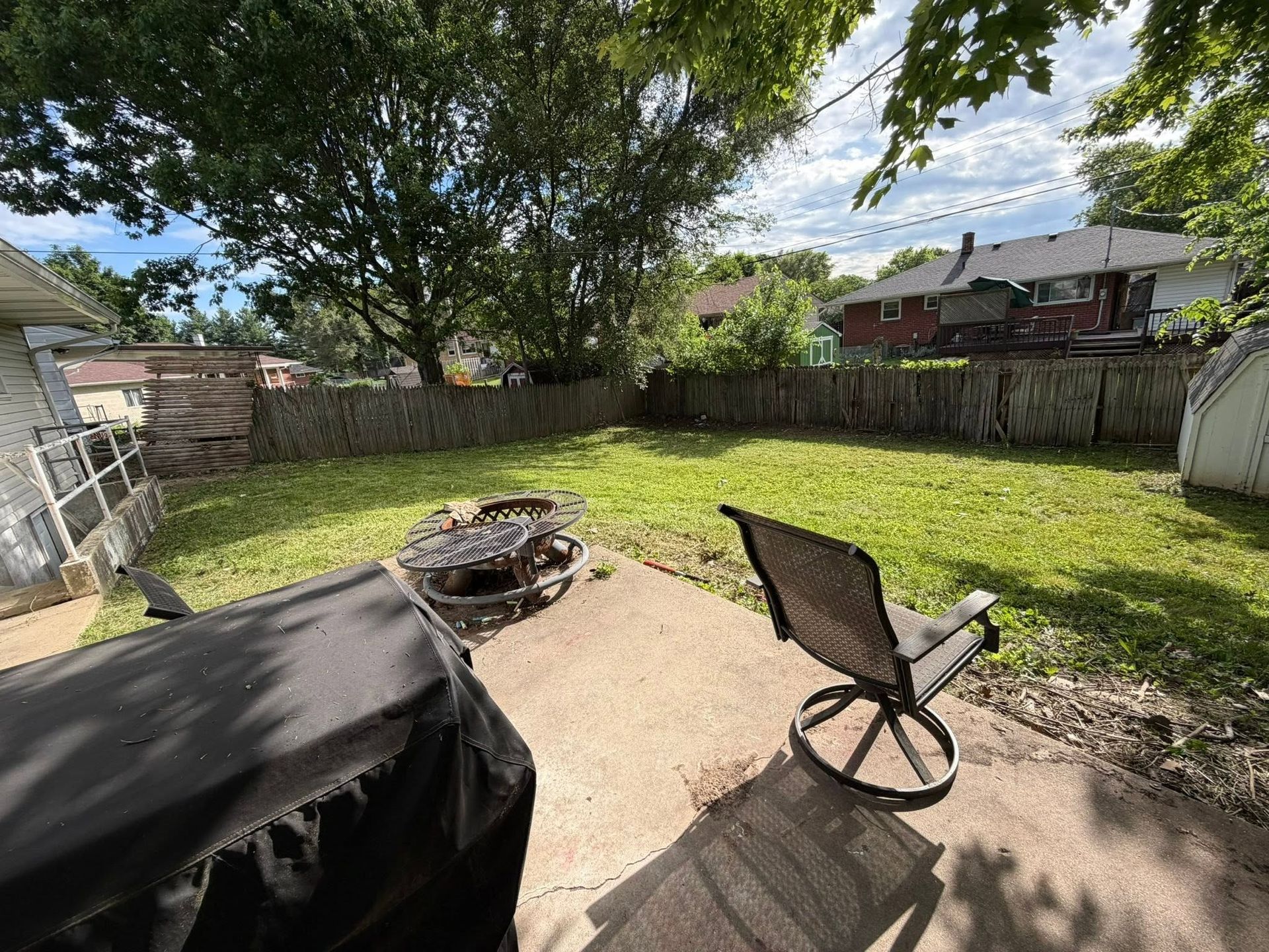 Backyard patio with fire pit, black chair, green lawn, fence, and trees under a partly cloudy sky